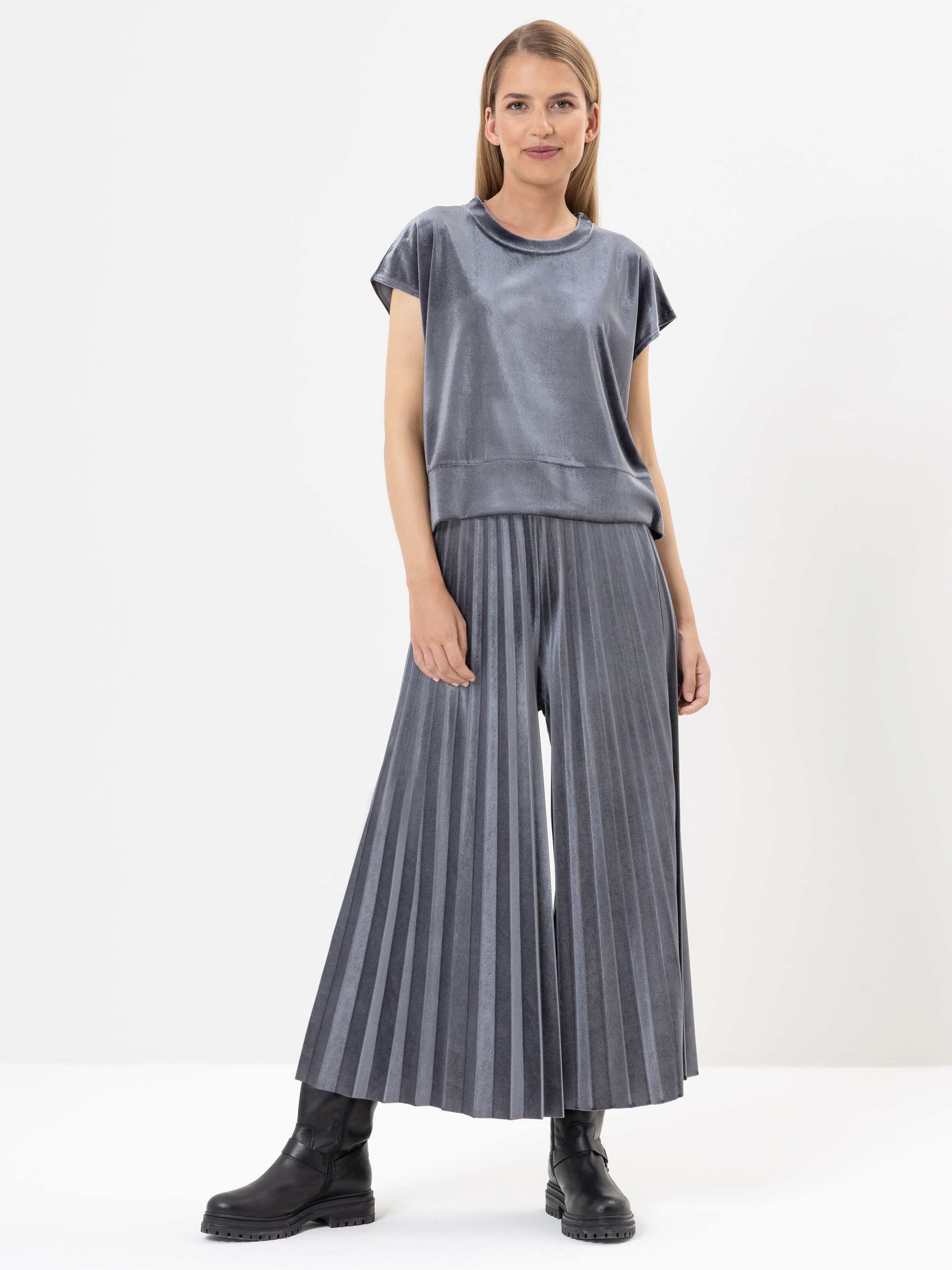 Eine Frau steht vor einem weißen Hintergrund. Sie trägt ein locker sitzendes, kurzärmeliges graues Oberteil und eine graue Hose mit weitem Bein, die in Falten gelegt ist, gepaart mit schwarzen Stiefeln. Sie hat glattes, hellbraunes Haar und einen neutr