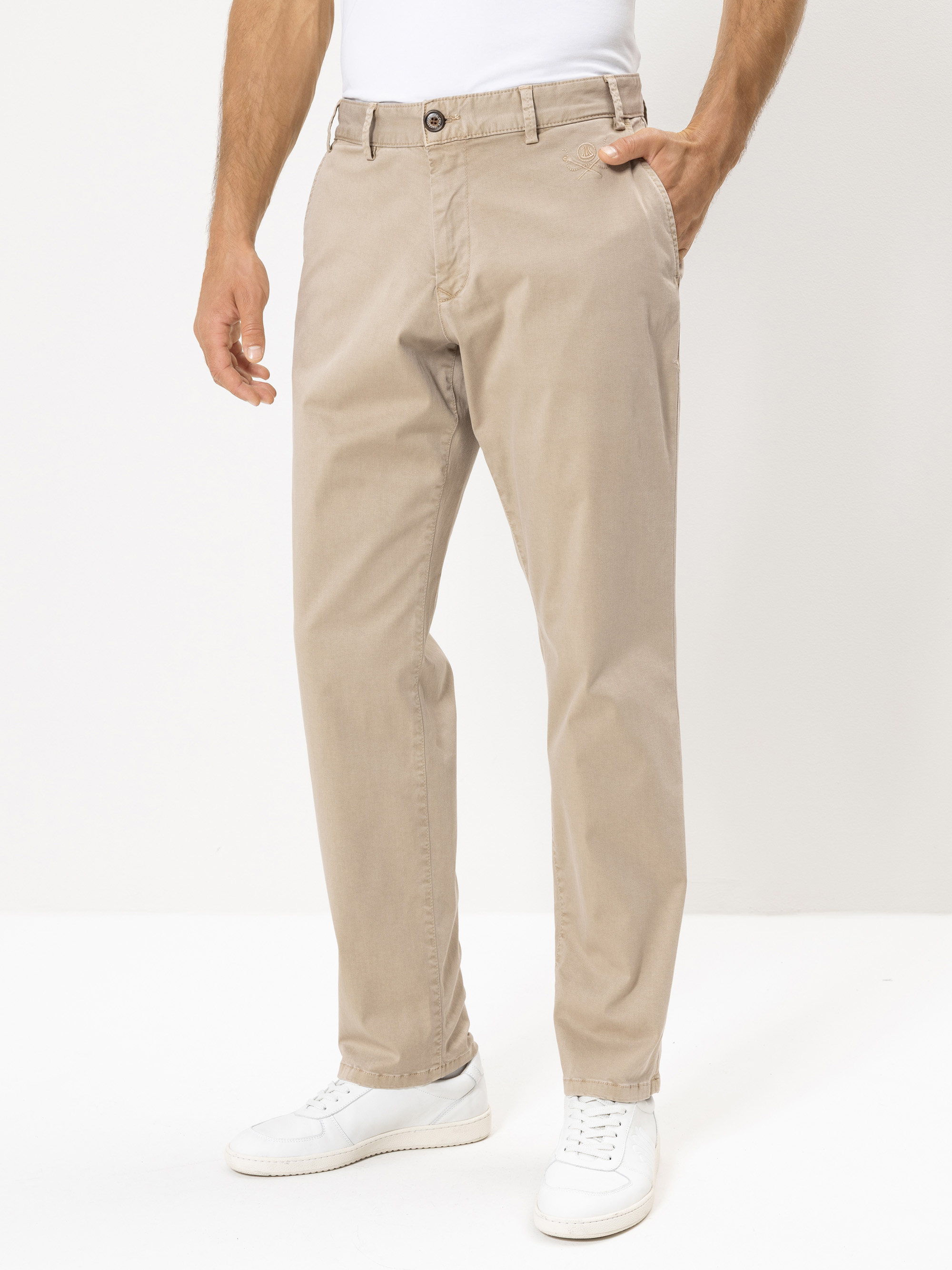 Ein Mann, der eine beigefarbene Chinohose mit der linken Hand in der Tasche, ein weißes T-Shirt und weiße Turnschuhe trägt, steht vor einem schlichten hellen Hintergrund.