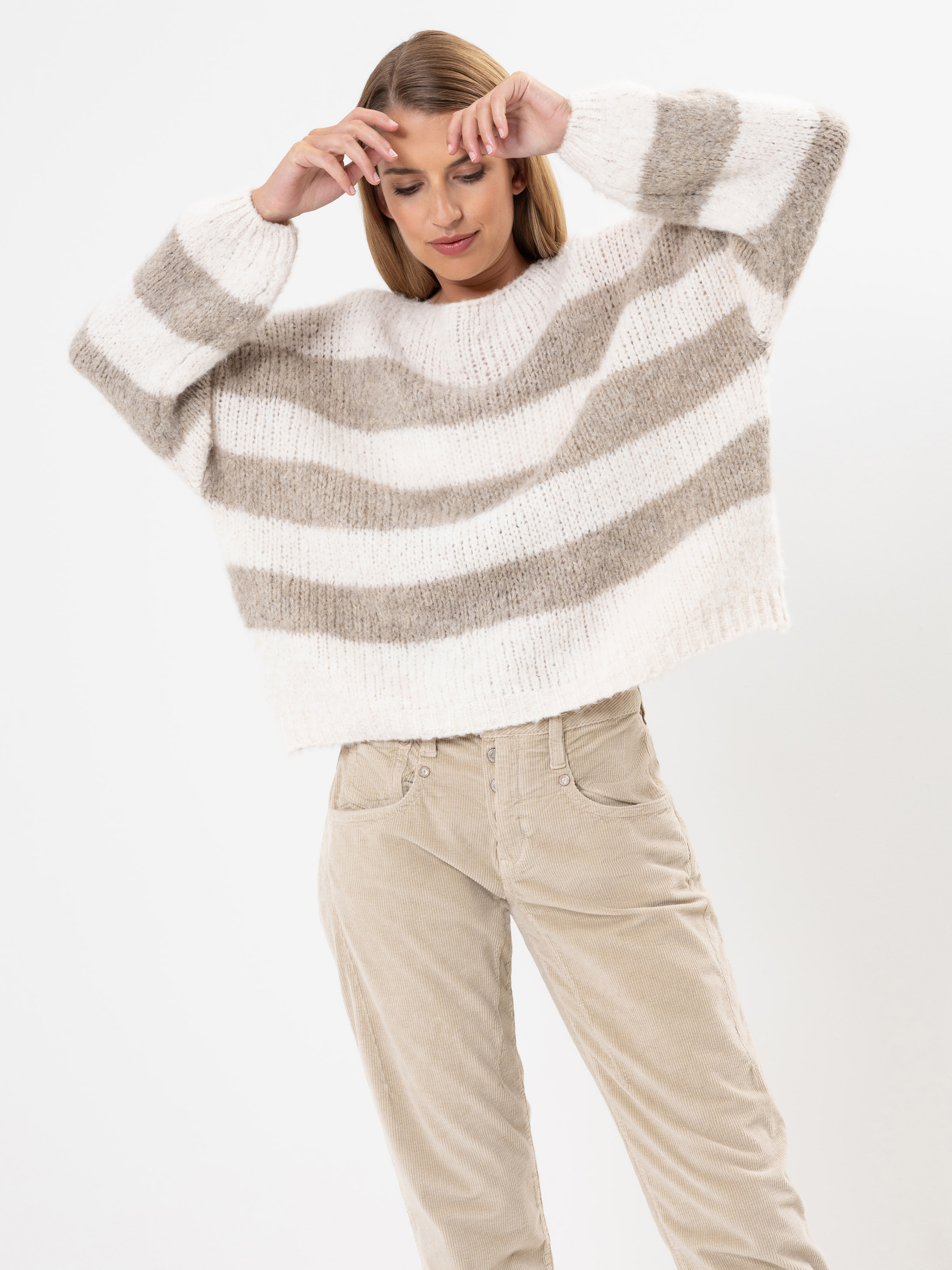Eine Frau, die einen lockeren, flauschigen Pullover mit breiten beigen und weißen Querstreifen und eine hellbeige Cordhose trägt, posiert mit erhobenen Armen vor einem weißen Hintergrund.