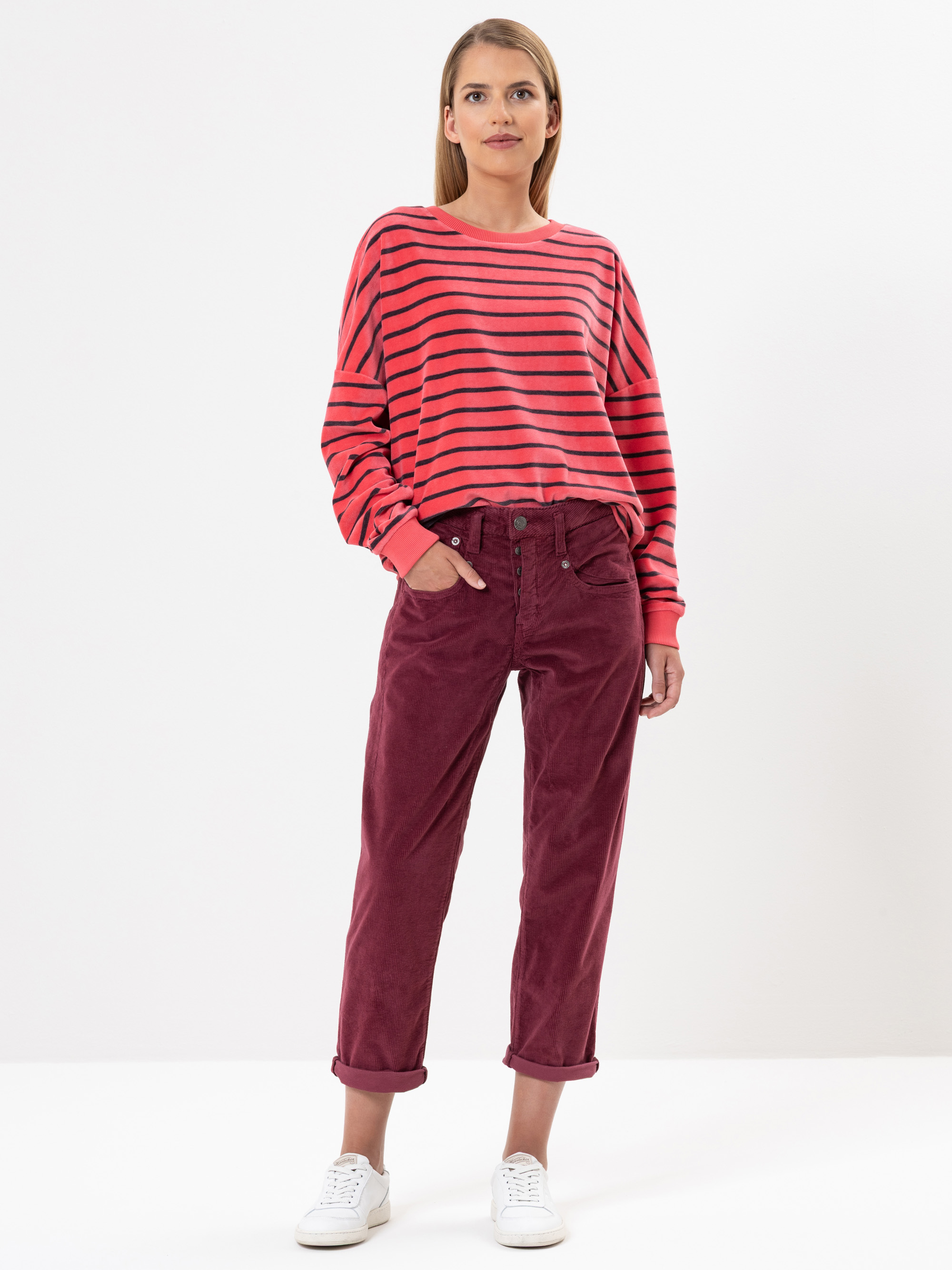 Eine Frau steht vor einem weißen Hintergrund und trägt ein rot-schwarz gestreiftes langärmeliges Hemd, eine kastanienbraune Cordhose mit aufgerollten Manschetten und weiße Turnschuhe. Sie hat glattes, schulterlanges Haar und eine entspannte Pose.