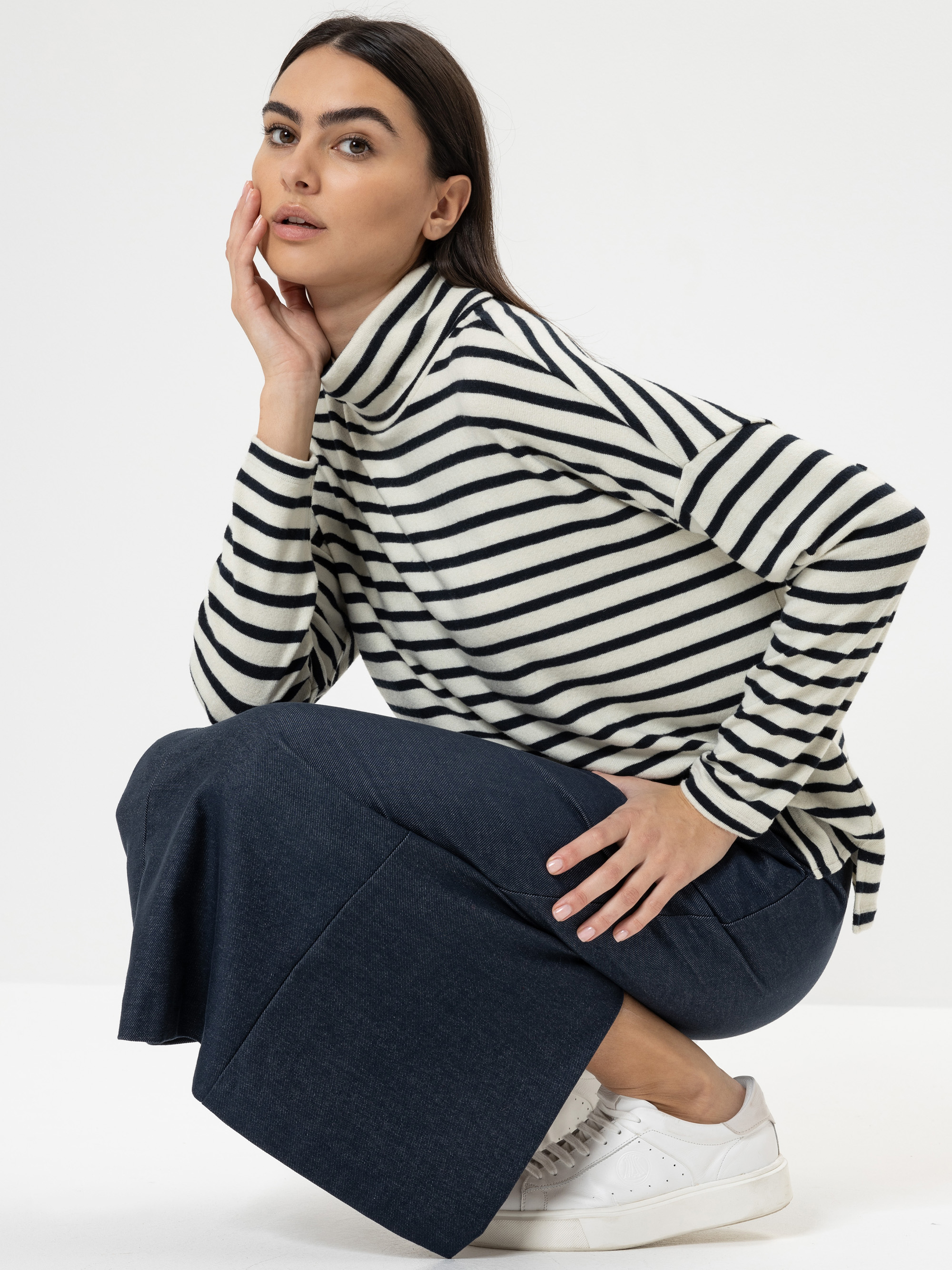 Eine Frau mit langen dunklen Haaren hockt auf dem Boden und stützt eine Hand auf ihre Wange. Sie trägt einen schwarz-weiß gestreiften Rollkragenpullover, einen langen dunklen Rock und weiße Turnschuhe vor einem schlichten weißen Hintergrund.