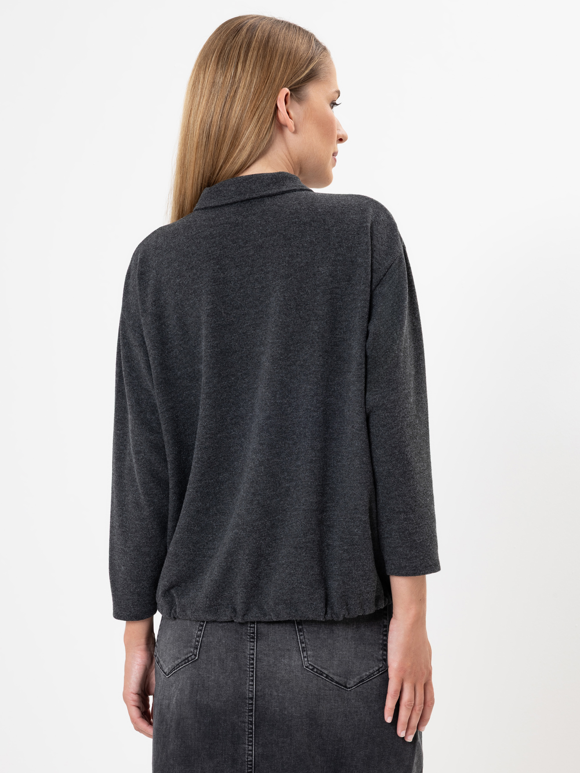 Eine Frau mit glattem, hellbraunem Haar wird von hinten gezeigt. Sie trägt ein dunkelgraues, langärmeliges Oberteil und einen verblichenen schwarzen Jeansrock vor einem schlichten weißen Hintergrund.
