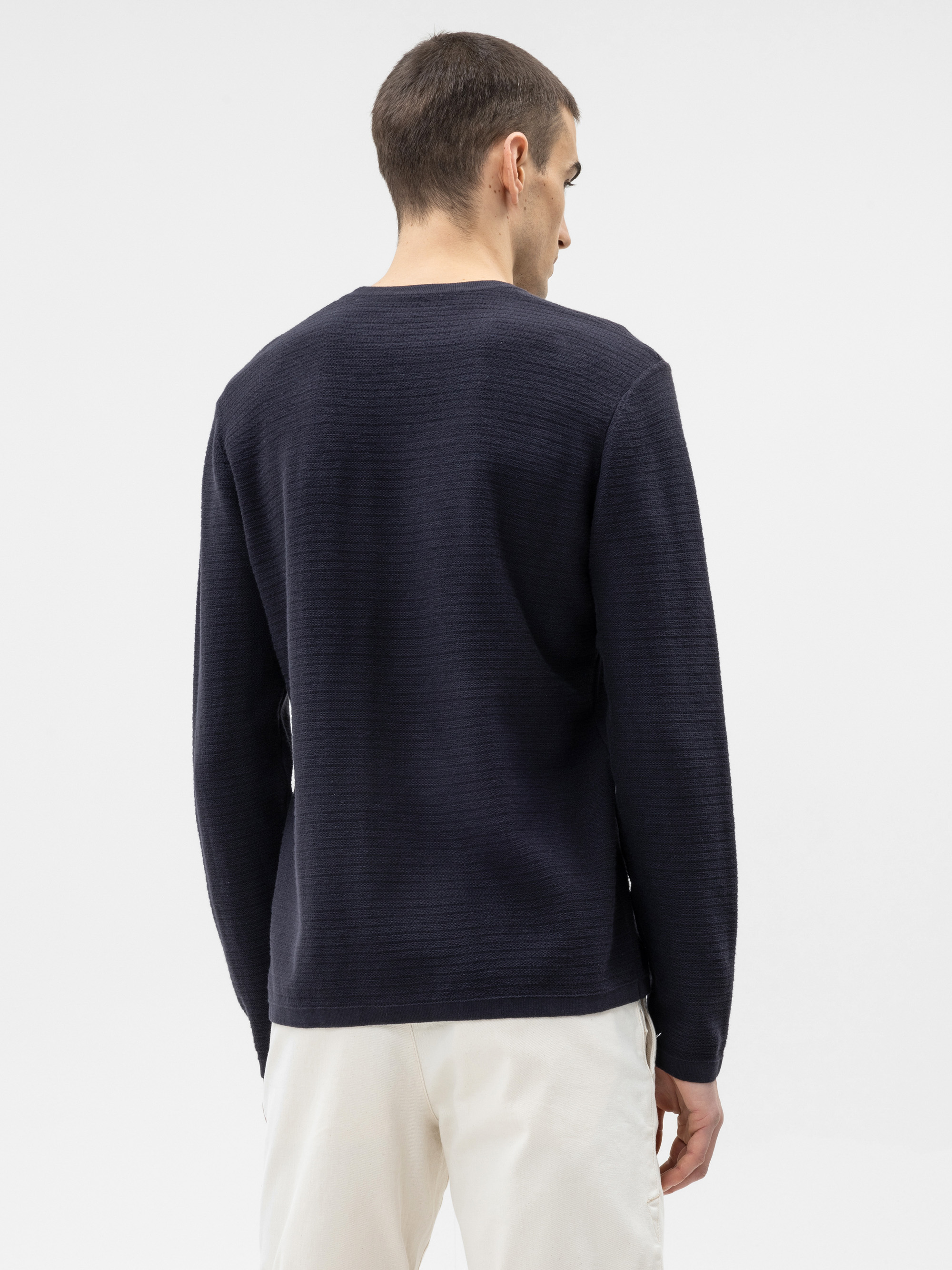 Ein Mann mit kurzen braunen Haaren, von hinten gesehen, trägt ein langärmeliges Hemd mit dunkler Struktur und eine helle Hose vor einem schlichten weißen Hintergrund.