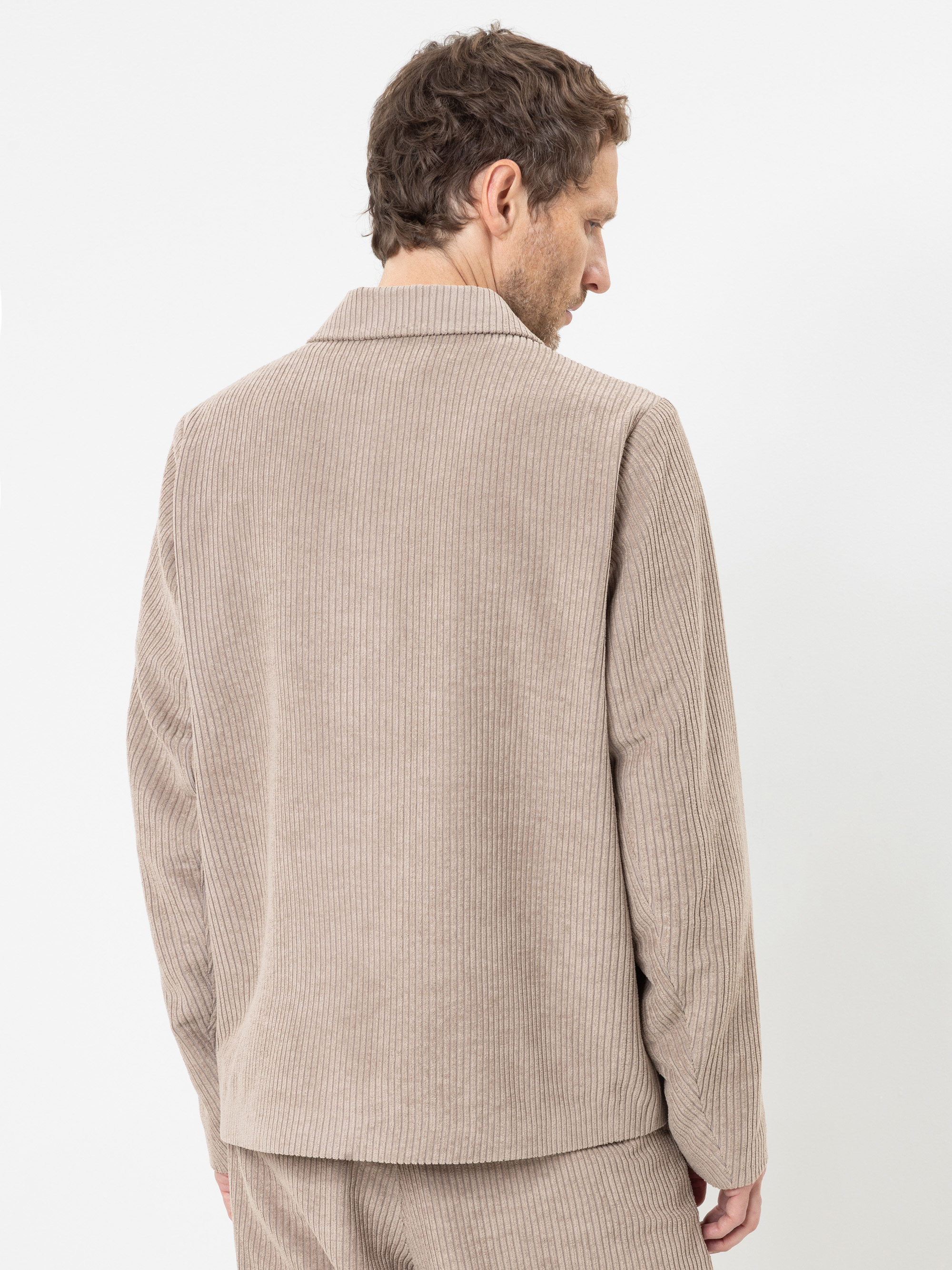 Vor einem schlichten weißen Hintergrund steht ein Mann mit hellbraunen Haaren, der mit dem Rücken zur Kamera steht und eine beige Cordjacke sowie eine dazu passende Hose trägt.