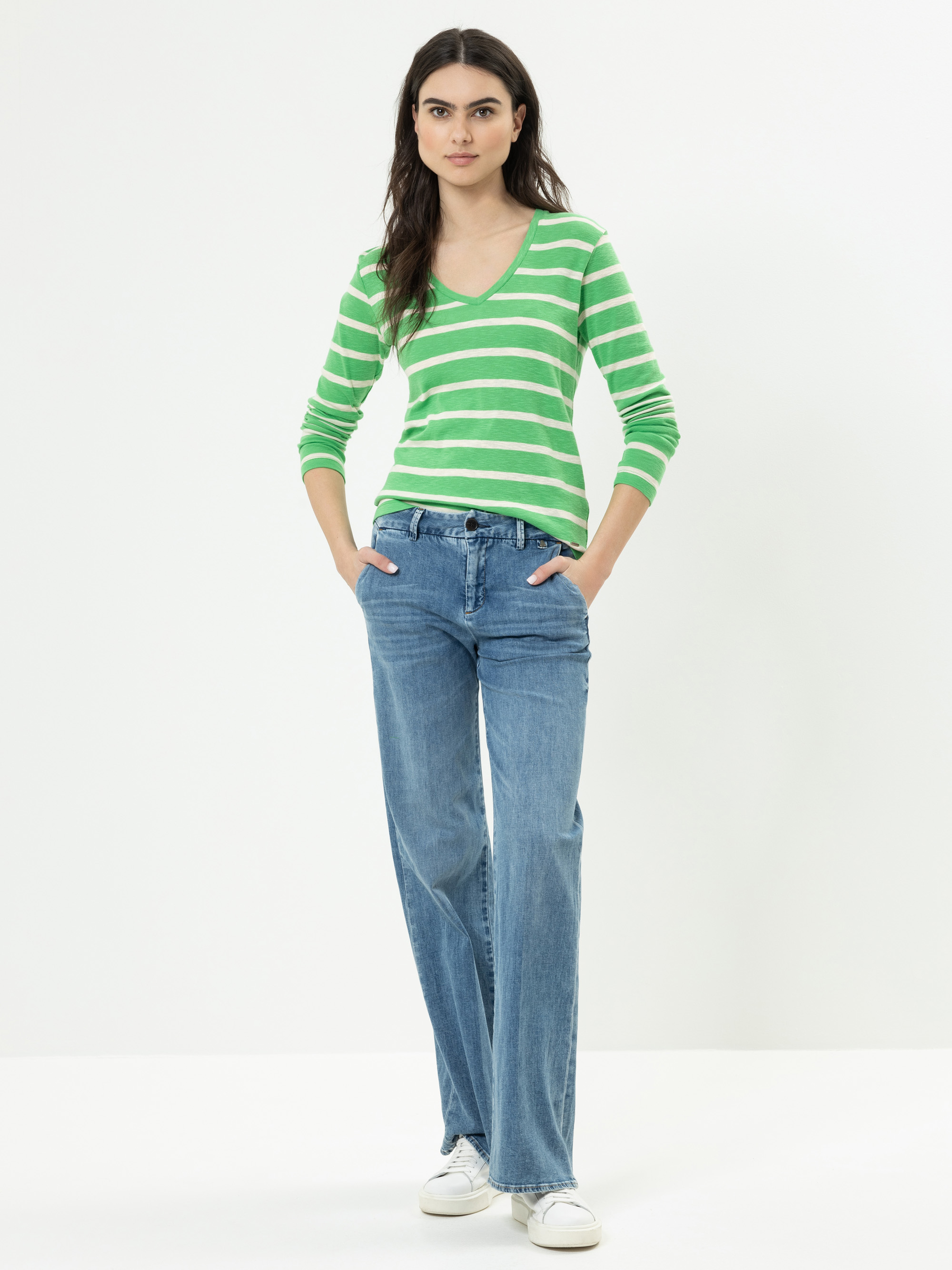 Eine Frau steht vor einem weißen Hintergrund und trägt ein grün-weiß gestreiftes langärmeliges Hemd, blaue Jeans mit weitem Bein und weiße Turnschuhe. Ihre Hände sind in den Taschen und sie schaut in die Kamera.
