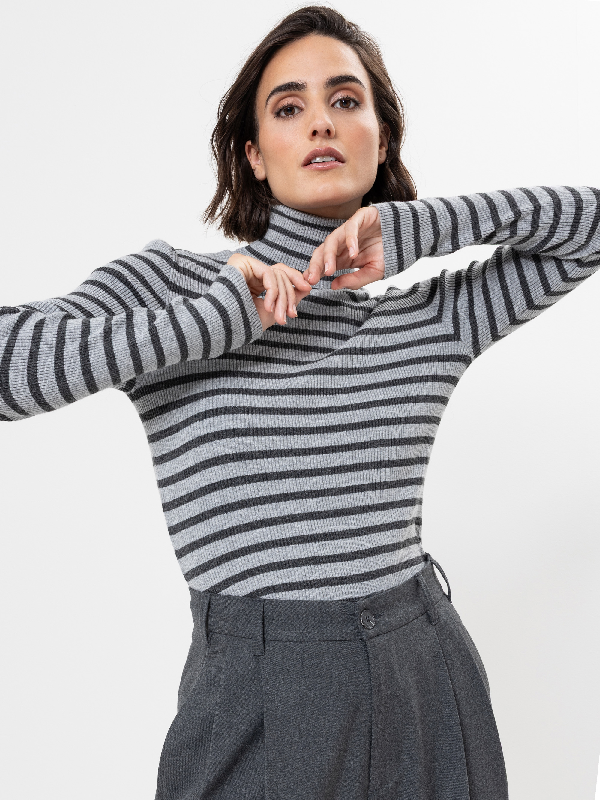 Eine Frau mit kurzen braunen Haaren trägt einen grau-schwarz gestreiften Rollkragenpullover und eine graue Hose mit hoher Taille. Sie posiert mit erhobenen Armen und verschränkten Fingern vor einem schlichten weißen Hintergrund.