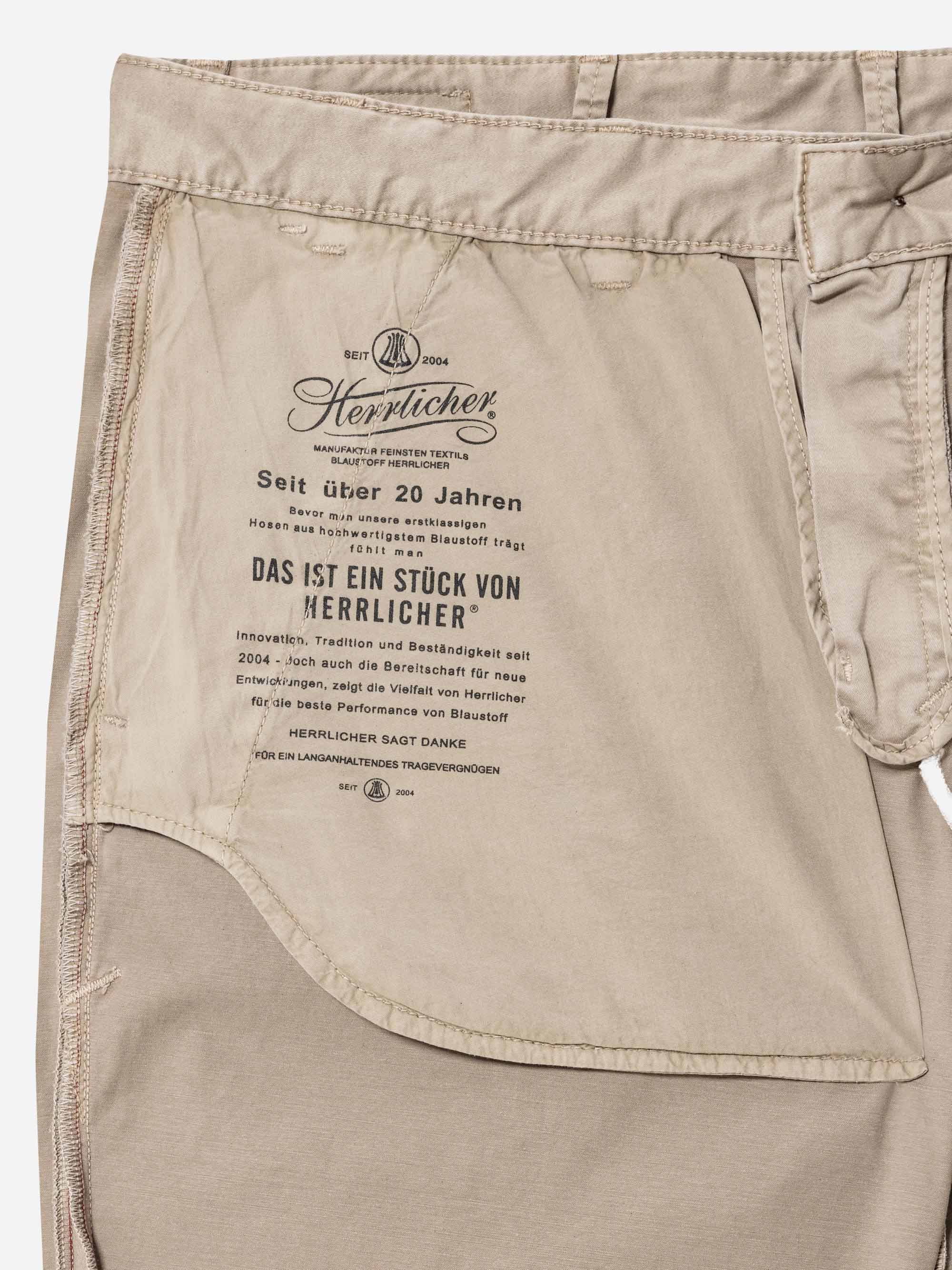 Nahaufnahme der Innentasche einer beigen Hose, die ein gedrucktes Etikett in deutscher Sprache mit dem Markennamen Herrlicher und Informationen über die Geschichte und Qualität des Unternehmens zeigt.