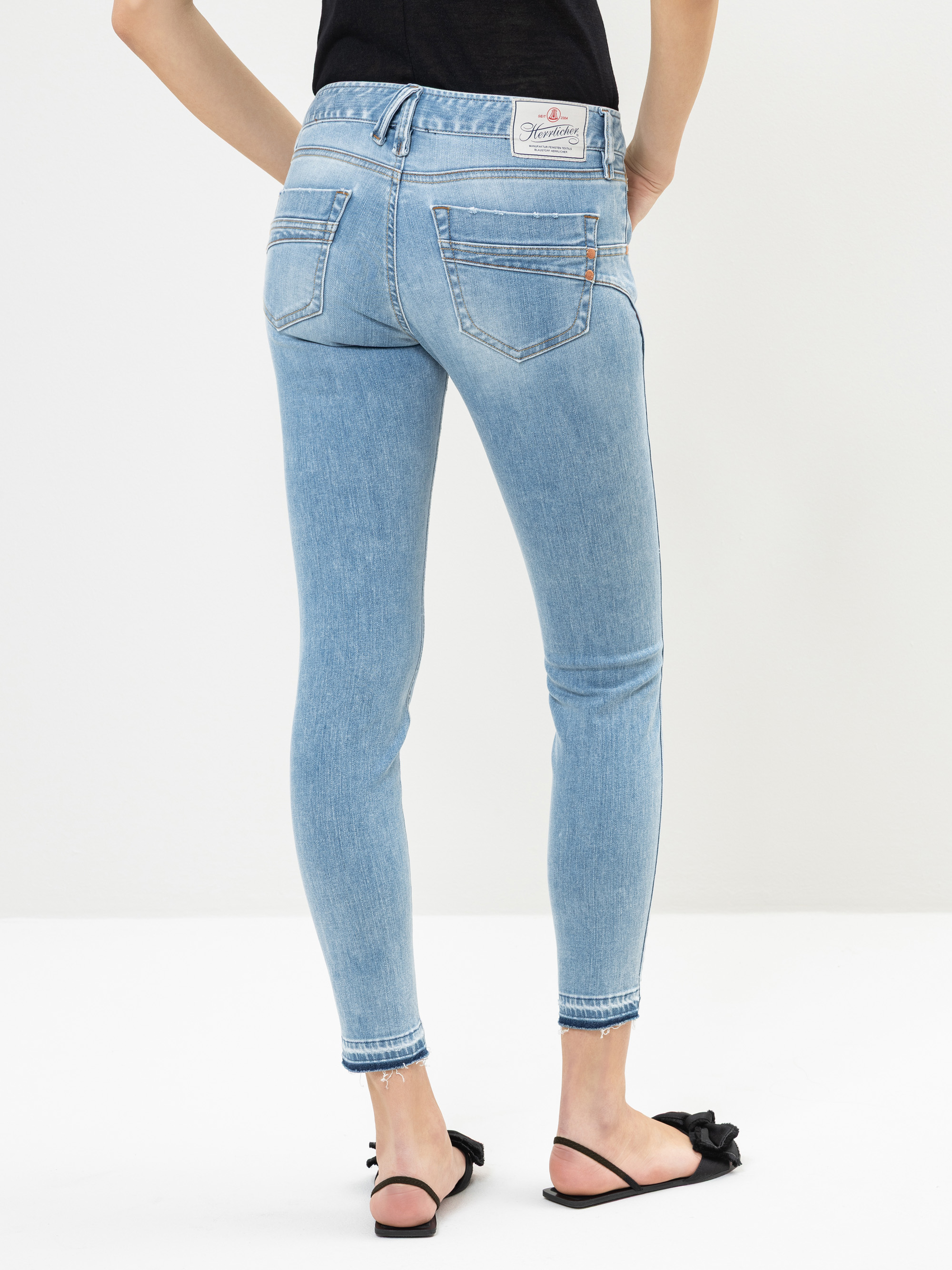 Eine Person in hellblauen Skinny-Jeans und schwarzen Sandalen steht mit dem Gesicht zur Kamera vor einem schlichten weißen Hintergrund. Ihre Hände sind in die Hüften gestemmt.