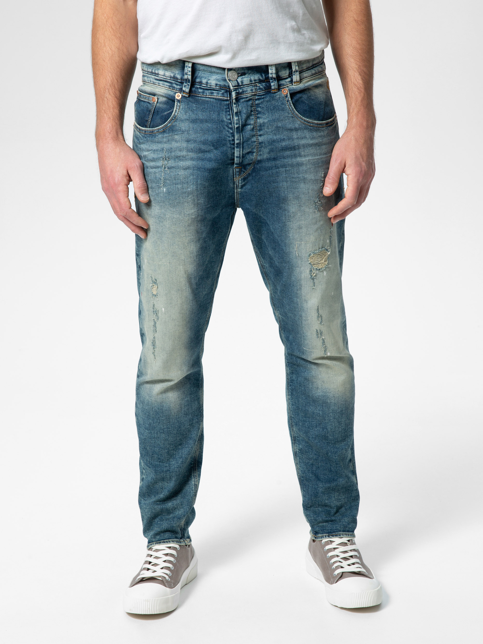 Eine Person, die hellblaue Jeans, ein weißes T-Shirt und graue Turnschuhe trägt, steht vor einem schlichten weißen Hintergrund. Die Jeans haben verblasste und zerrissene Details auf der Vorderseite.