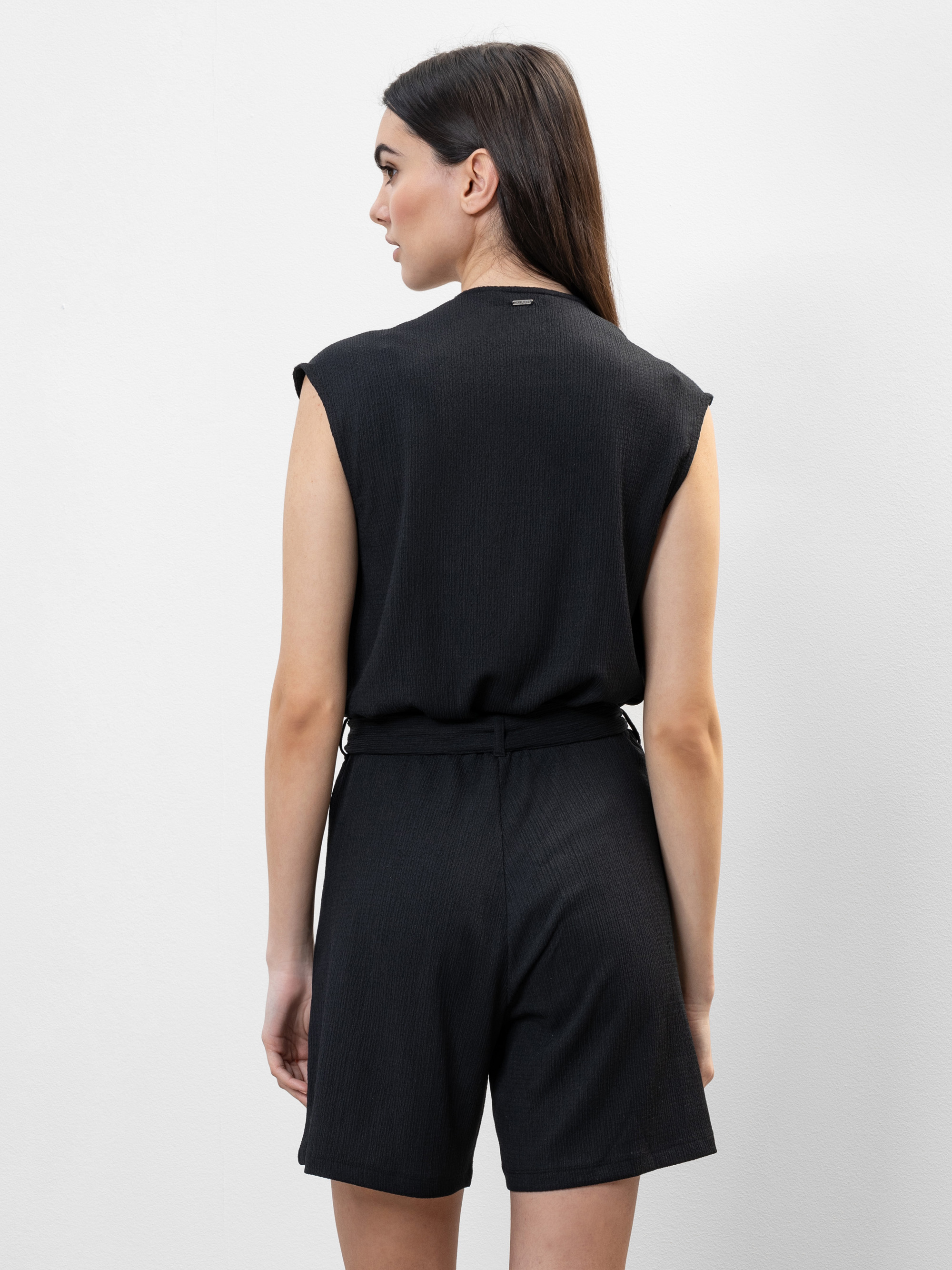 Eine Frau mit langen braunen Haaren steht mit dem Gesicht zur Wand und trägt einen ärmellosen schwarzen Strampler mit Gürtel in der Taille vor einem schlichten weißen Hintergrund.