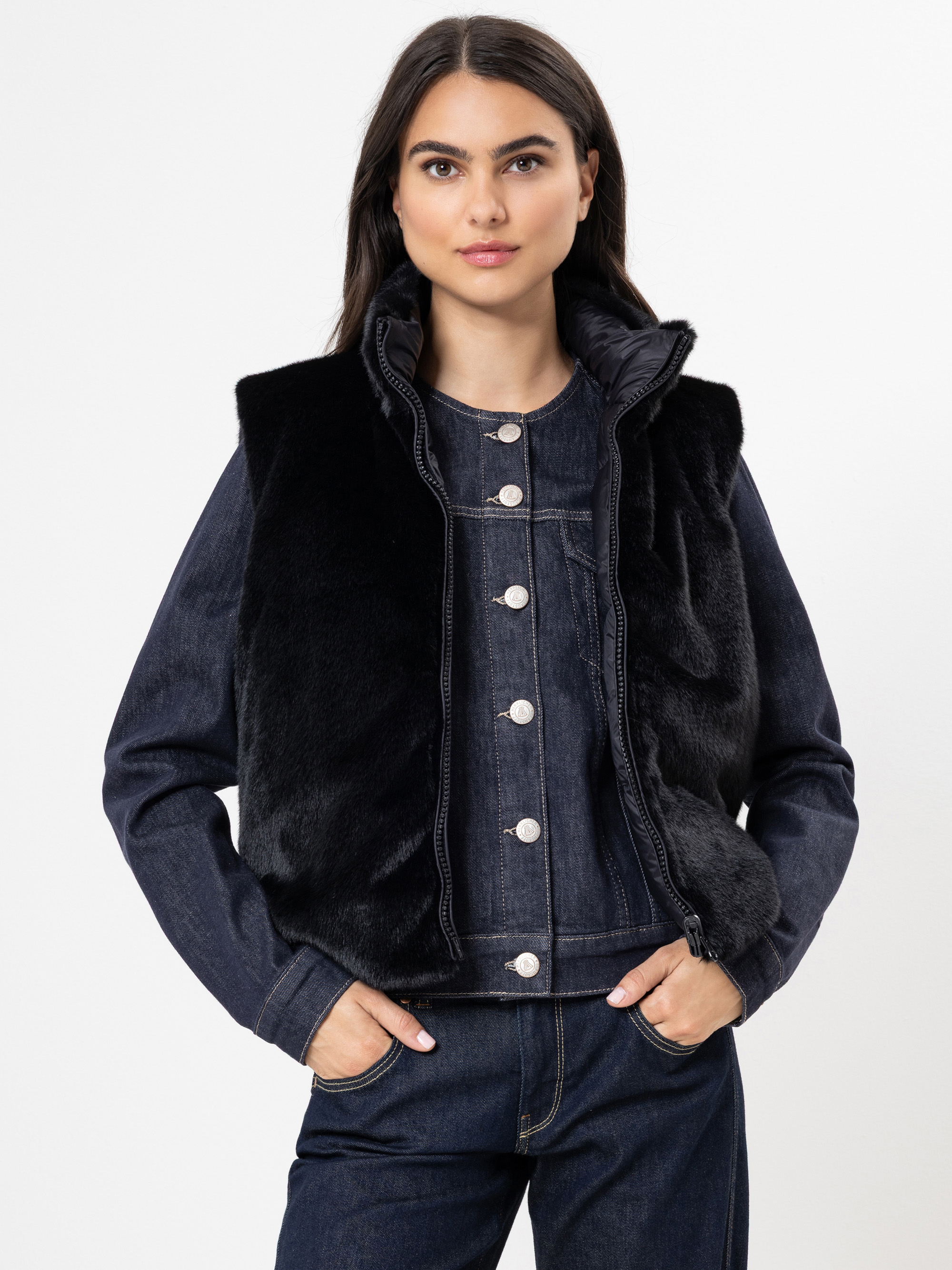 Eine Frau mit langen dunklen Haaren trägt eine schwarze Kunstpelzweste über einer dunklen Jeansjacke mit silbernen Knöpfen und dazu passende Jeans, die vor einem schlichten weißen Hintergrund stehen.