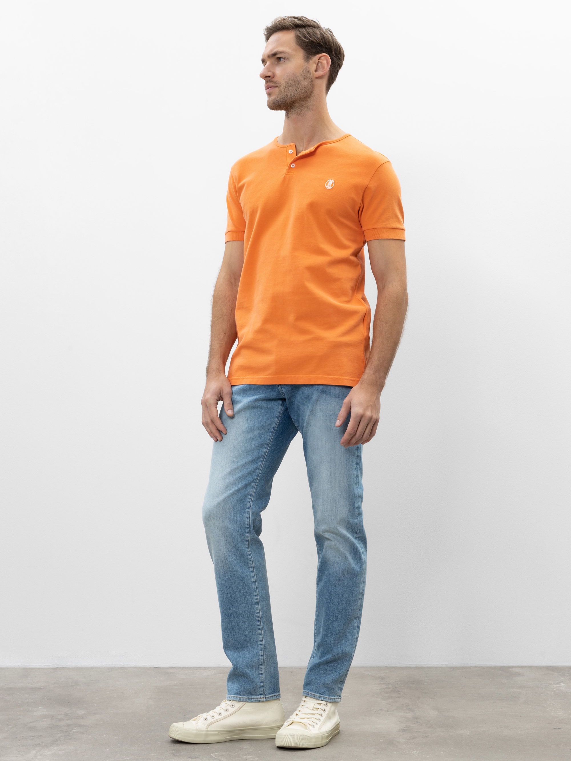 Ein Mann steht vor einem schlichten weißen Hintergrund und trägt ein leuchtend orangefarbenes Poloshirt, hellblaue Jeans und weiße Turnschuhe. Er blickt leicht nach rechts, eine Hand an der Seite, die andere am Oberschenkel haltend.