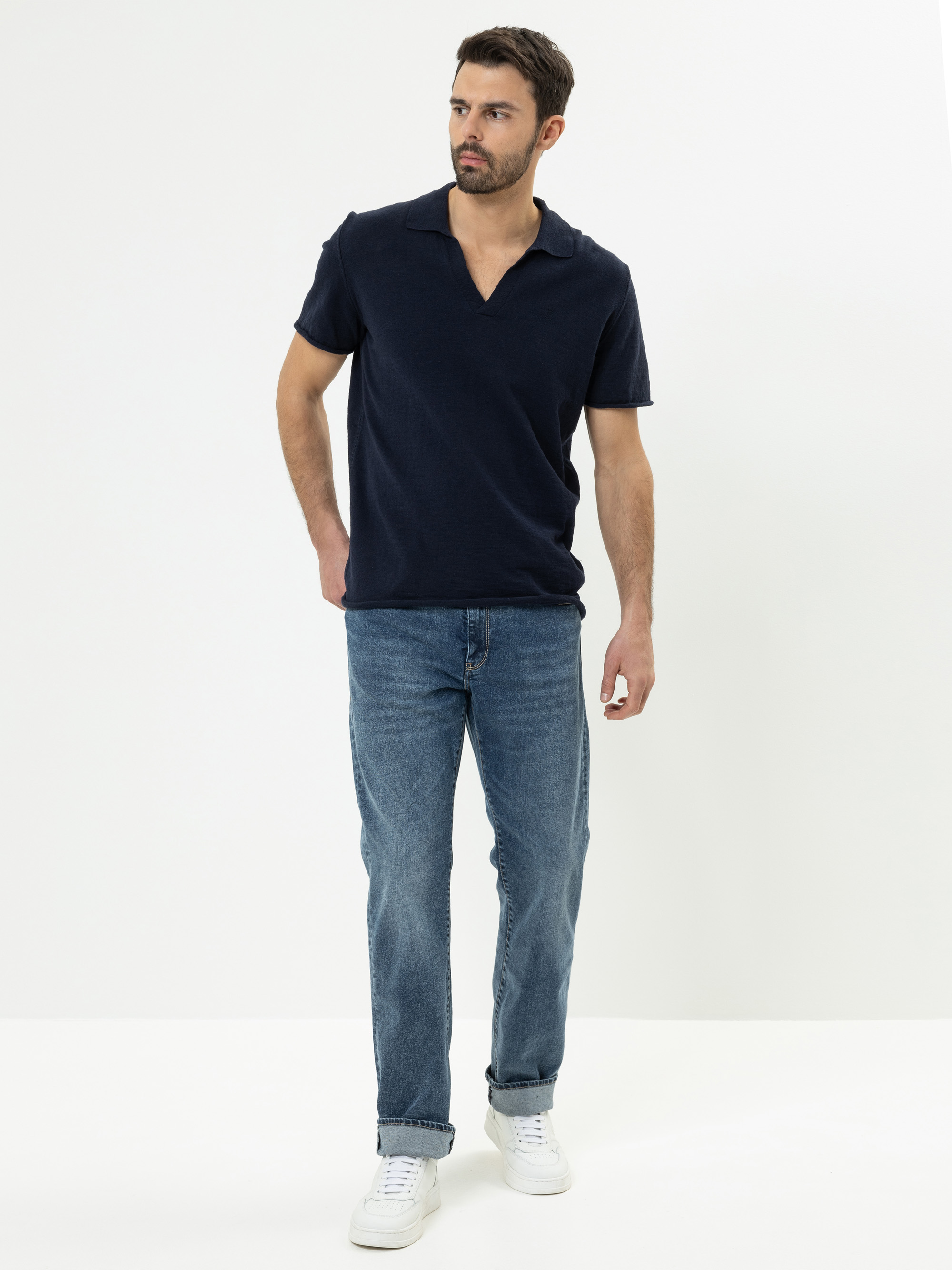 Ein Mann mit kurzen dunklen Haaren und einem Bart steht vor einem schlichten weißen Hintergrund. Er trägt ein marineblaues, kurzärmeliges Poloshirt, blaue Jeans und weiße Turnschuhe. Er blickt leicht nach links und hält eine Hand hinter dem Rücken.