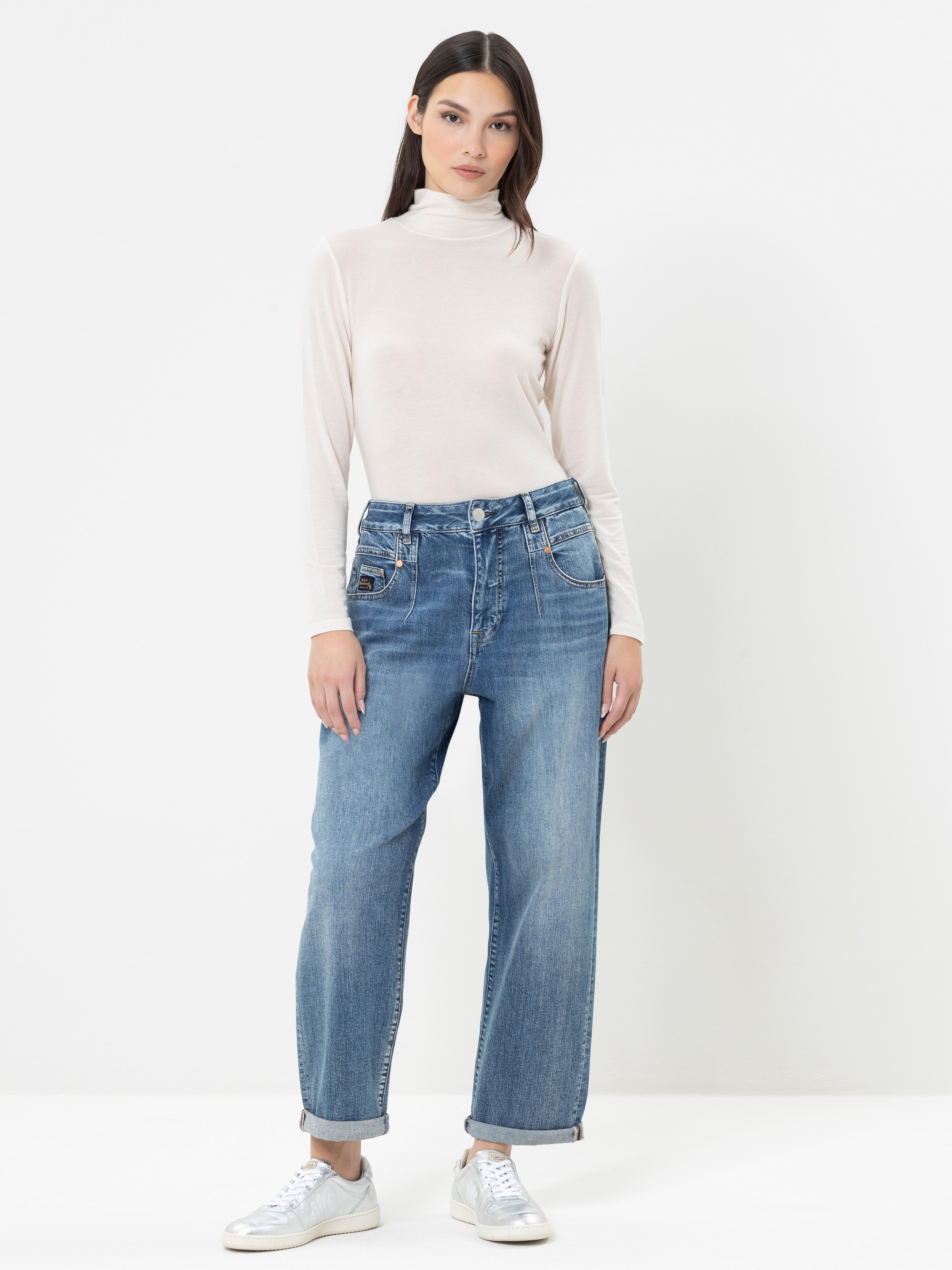 Eine Frau steht vor einem schlichten weißen Hintergrund und trägt einen durchsichtigen weißen Rollkragenpullover, eine blaue Jeans mit hoher Taille und umgeschlagenen Bündchen sowie weiße Sneaker. Sie hat glattes dunkles Haar und einen neutralen Gesi