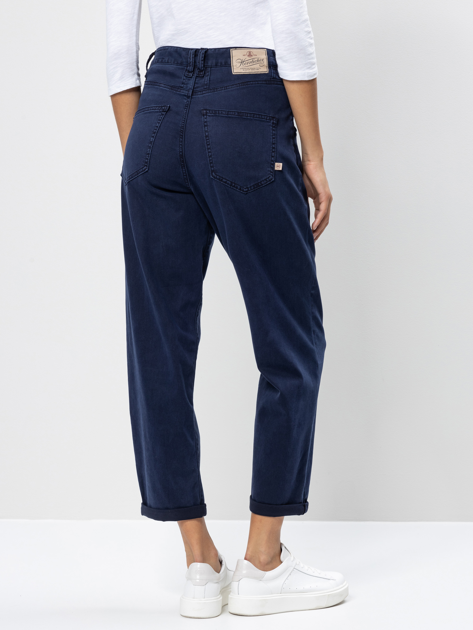 Person, die dunkelblaue Jeans mit hoher Taille und Bündchen am Saum, ein weißes langärmeliges Oberteil und weiße Turnschuhe trägt und vor einem schlichten hellen Hintergrund von der Kamera abgewandt steht.