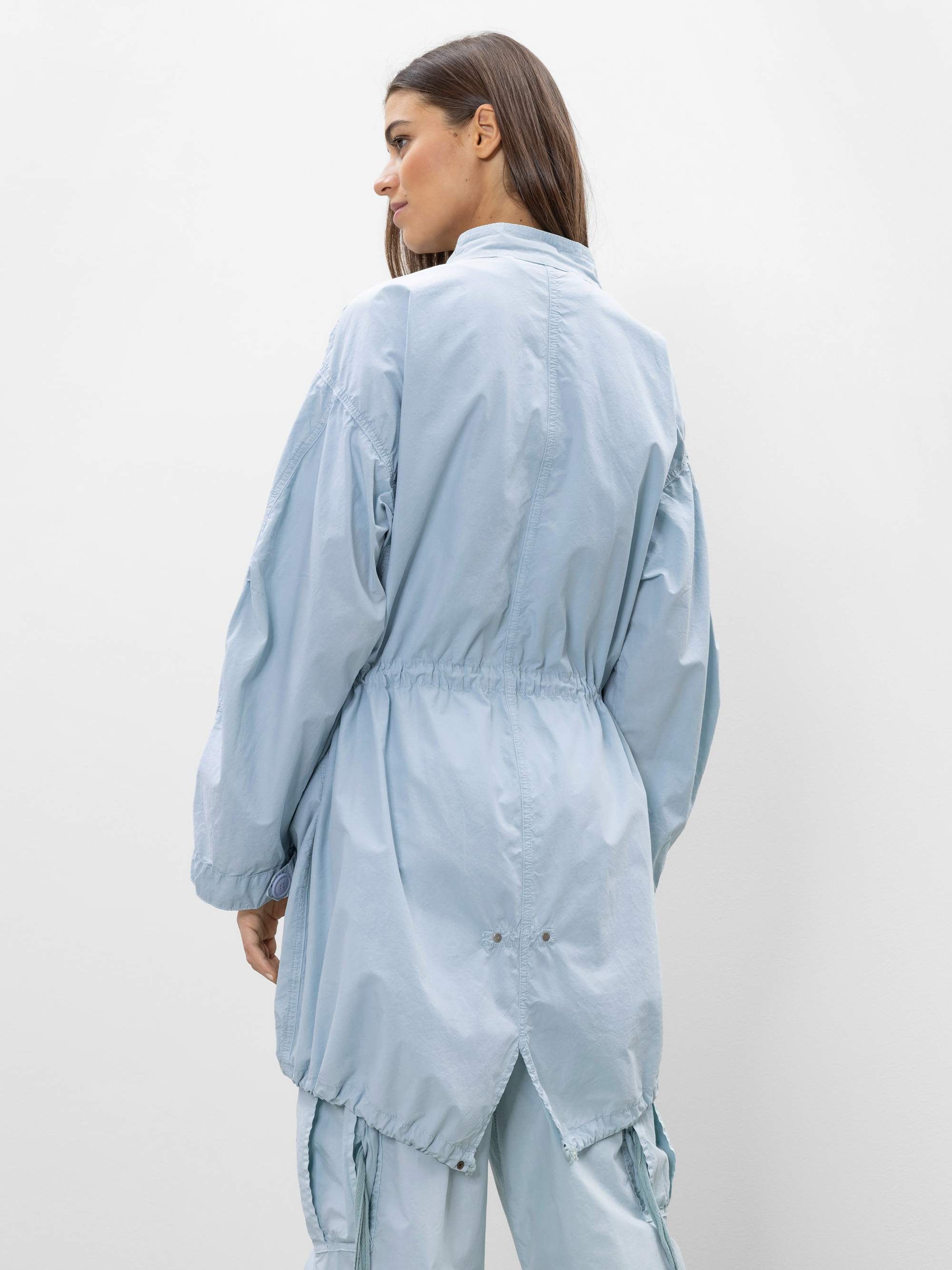 Eine Frau mit langen braunen Haaren trägt eine lockere, hellblaue Jacke und eine dazu passende Hose. Sie steht mit dem Rücken zur Kamera und blickt über ihre Schulter vor einem schlichten weißen Hintergrund.