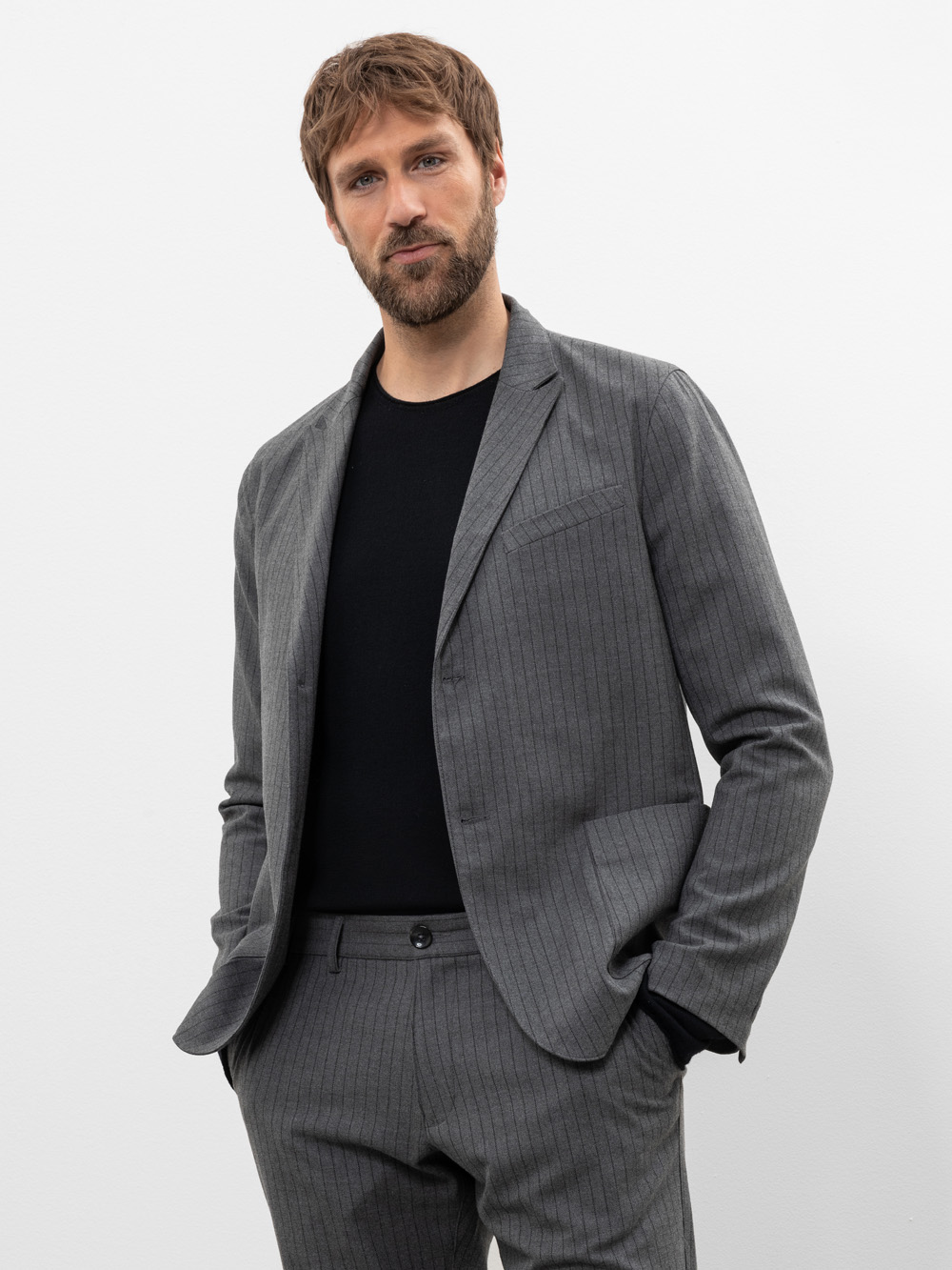Ein Mann mit kurzen braunen Haaren und einem Bart steht vor einem weißen Hintergrund, trägt einen grauen Nadelstreifenanzug und ein schwarzes Hemd, hat die Hände in den Taschen und blickt selbstbewusst in die Kamera.