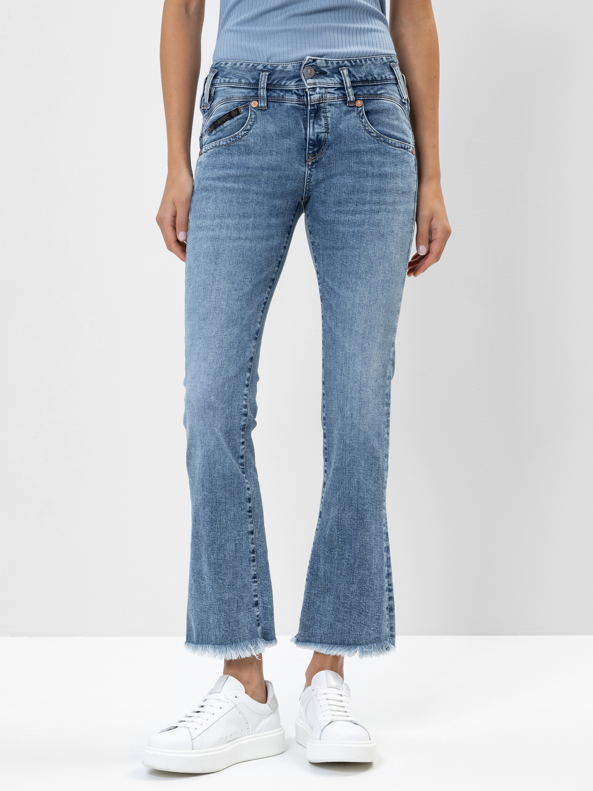 Eine Person, die hellblaue Schlagjeans mit ausgefransten Säumen, ein blaues geripptes Oberteil und weiße Turnschuhe trägt, steht vor einem schlichten weißen Hintergrund. Das Bild zeigt die Person von den Schultern bis zu den Füßen.