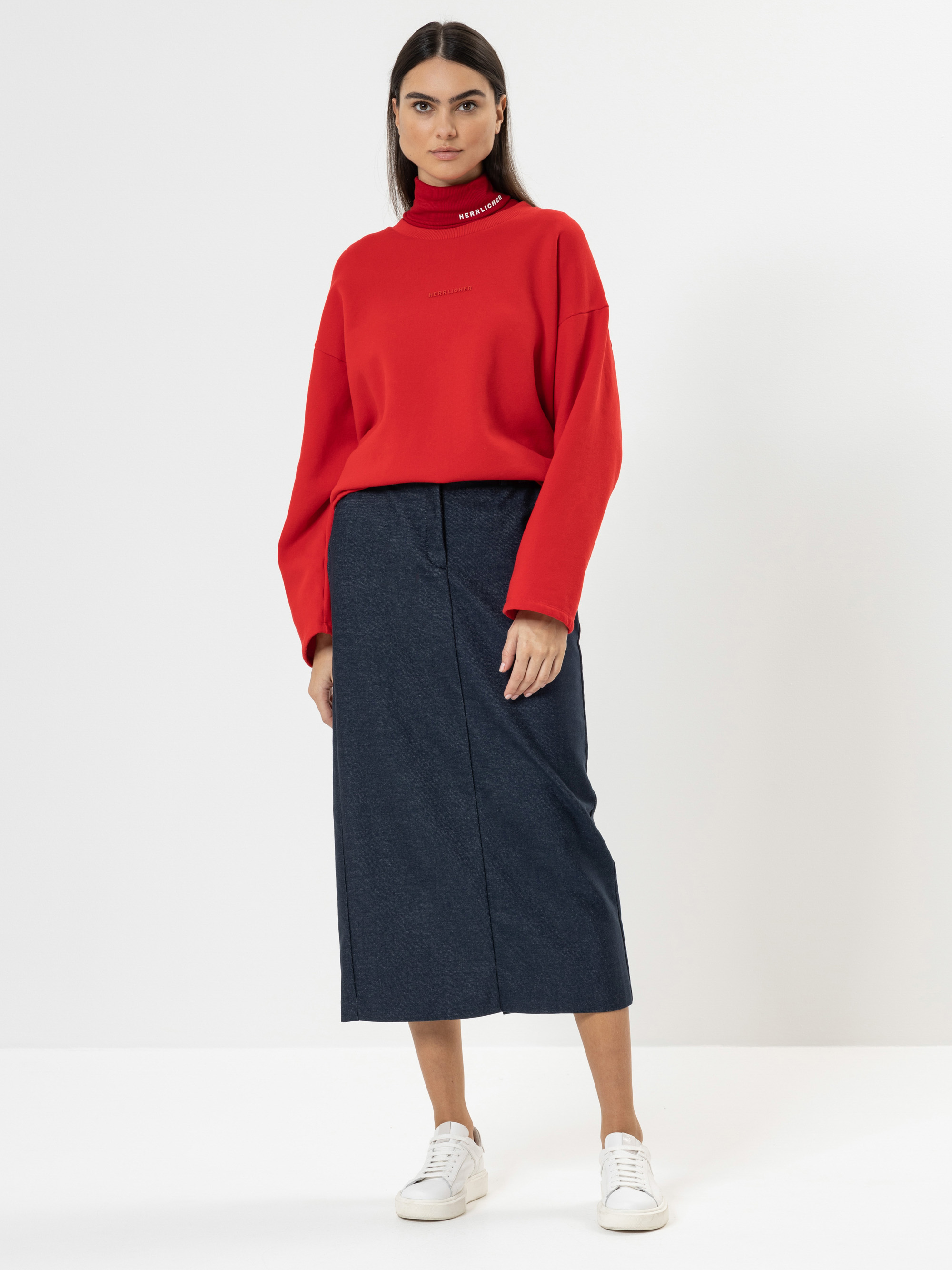 Eine Frau steht vor einem schlichten weißen Hintergrund und trägt einen roten Rollkragenpullover, einen langen dunkelblauen Rock und weiße Turnschuhe. Sie hat glattes dunkles Haar und einen neutralen Gesichtsausdruck.