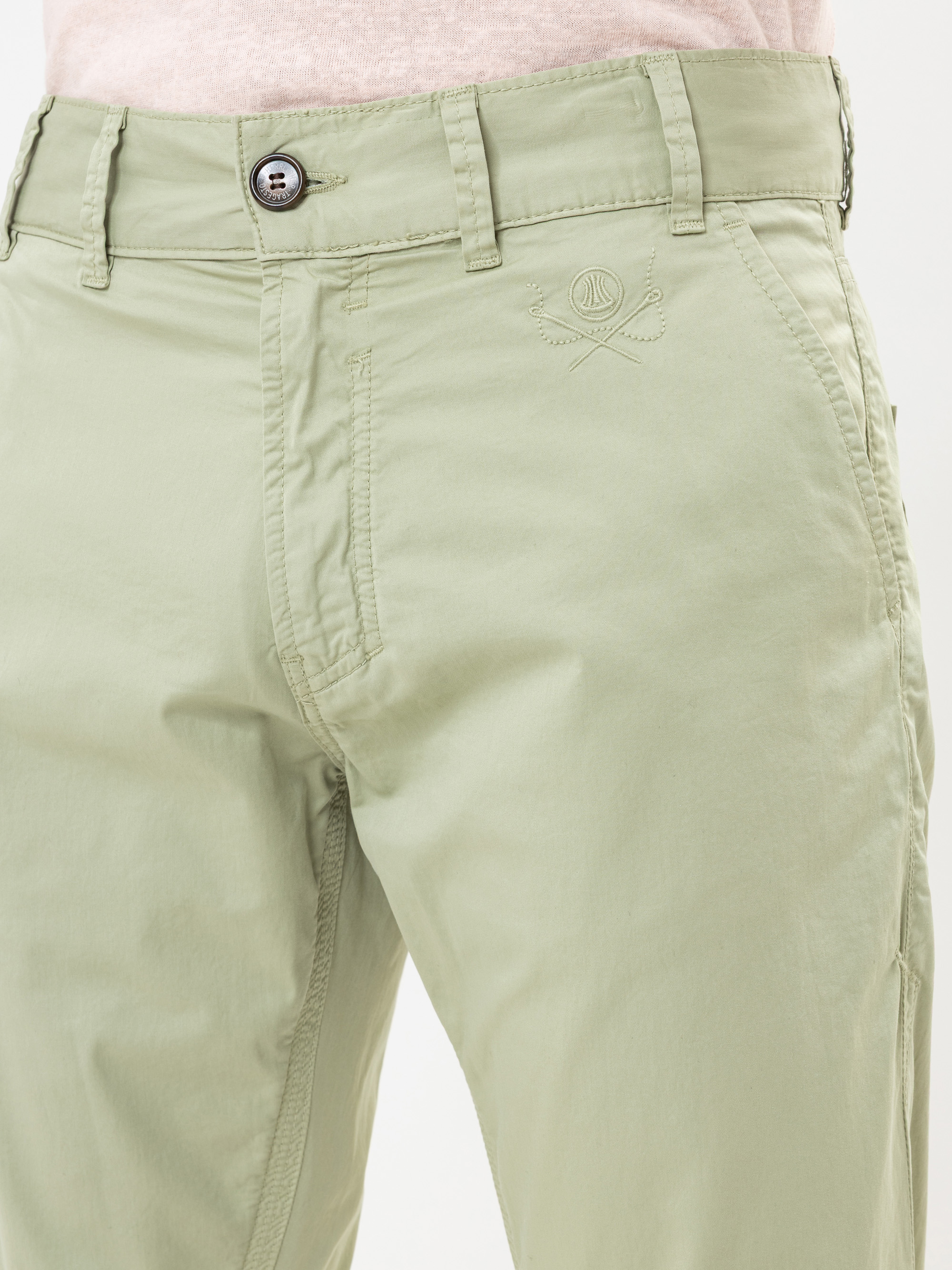 Nahaufnahme einer Person, die eine hellgrüne Hose mit Knopf und Reißverschluss, Gürtelschlaufen und einem gestickten Logo in der Nähe der Vordertasche trägt. Der Bund und die Oberschenkel sind sichtbar.