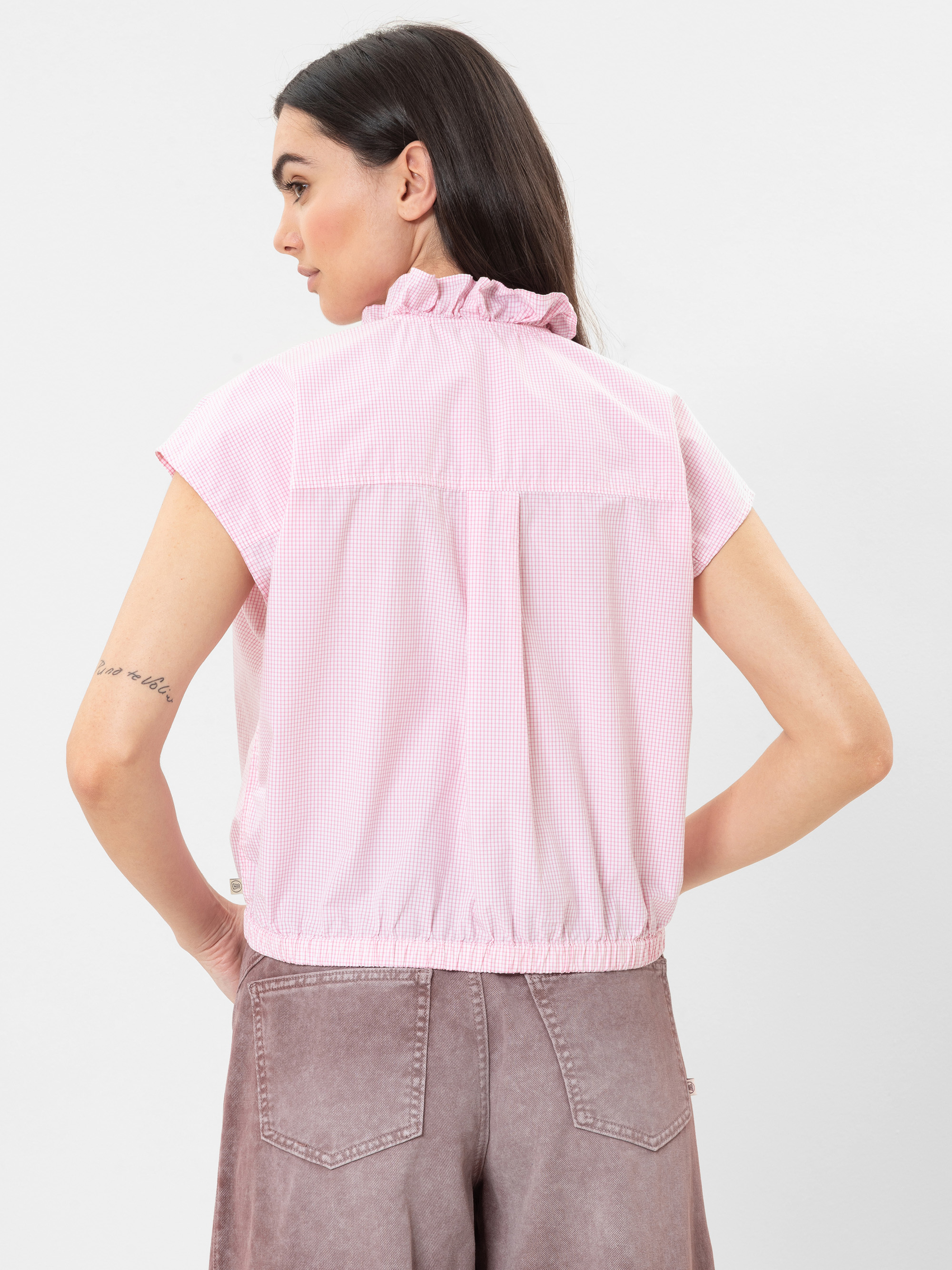 Eine Frau mit langen dunklen Haaren trägt eine hellrosa, kurzärmelige Bluse mit Rüschenkragen und lockerem Sitz, gepaart mit einer malvenfarbenen Hose mit hoher Taille, und steht mit dem Rücken zur Kamera.