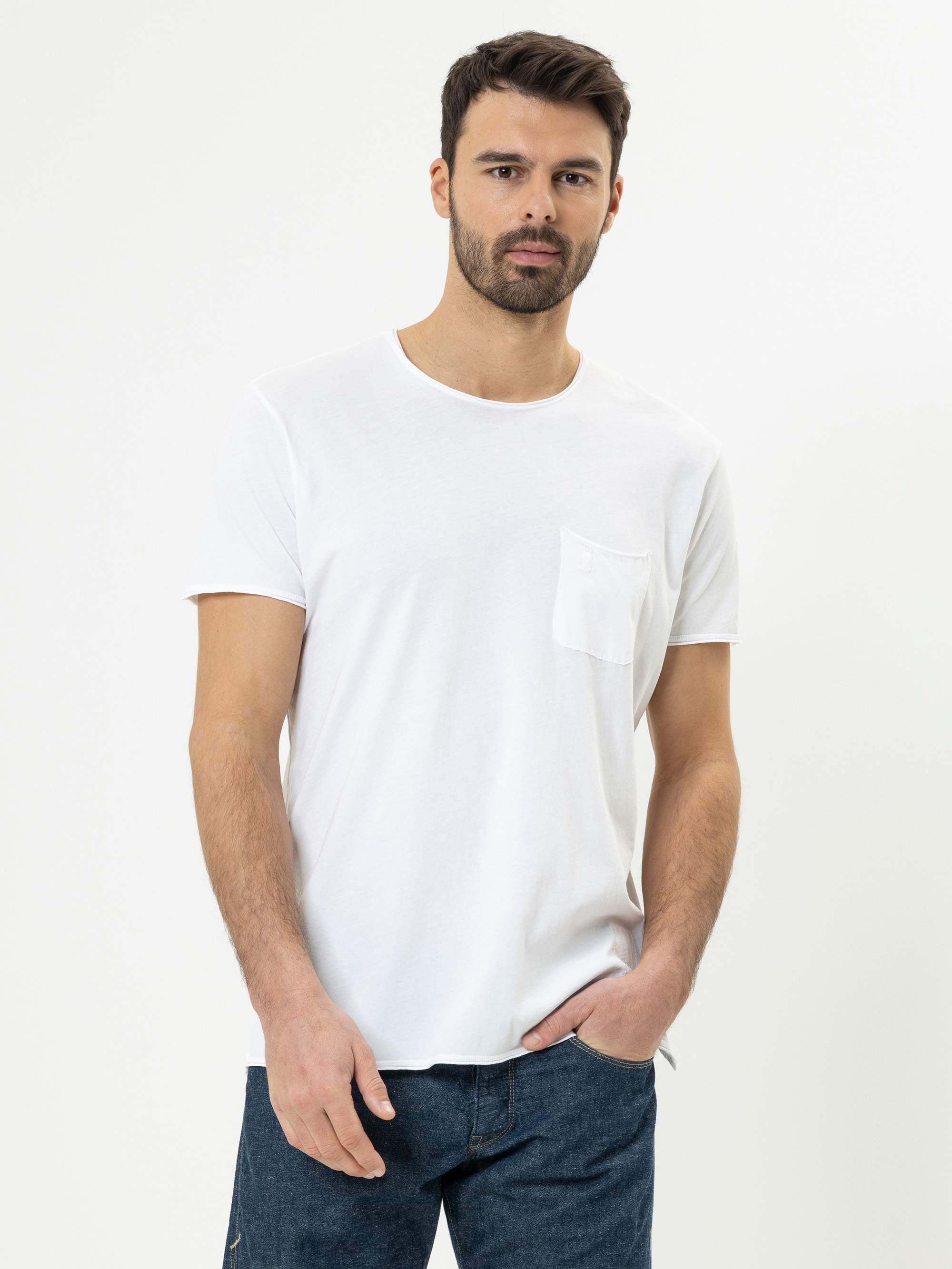 Ein Mann mit kurzen dunklen Haaren und Bart trägt ein schlichtes weißes T-Shirt mit Brusttasche und eine blaue Jeans und steht vor einem schlichten weißen Hintergrund.