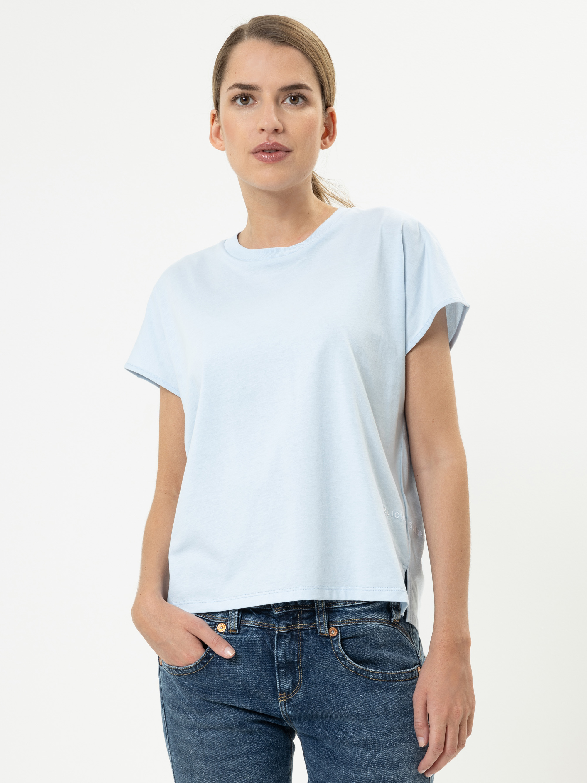 Eine Frau, die ein hellblaues, kurzärmeliges T-Shirt und blaue Jeans trägt, steht vor einem schlichten weißen Hintergrund, eine Hand in der Tasche, und schaut nach vorn.