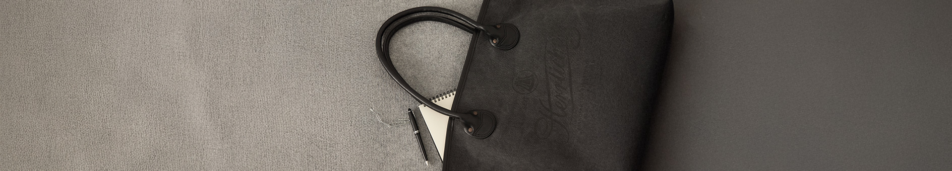 Eine schwarze Handtasche mit geschwungenen Griffen liegt auf einer hellgrauen Fläche und ist teilweise geöffnet, so dass ein weißes Notizbuch und ein silberner Stift zum Vorschein kommen.