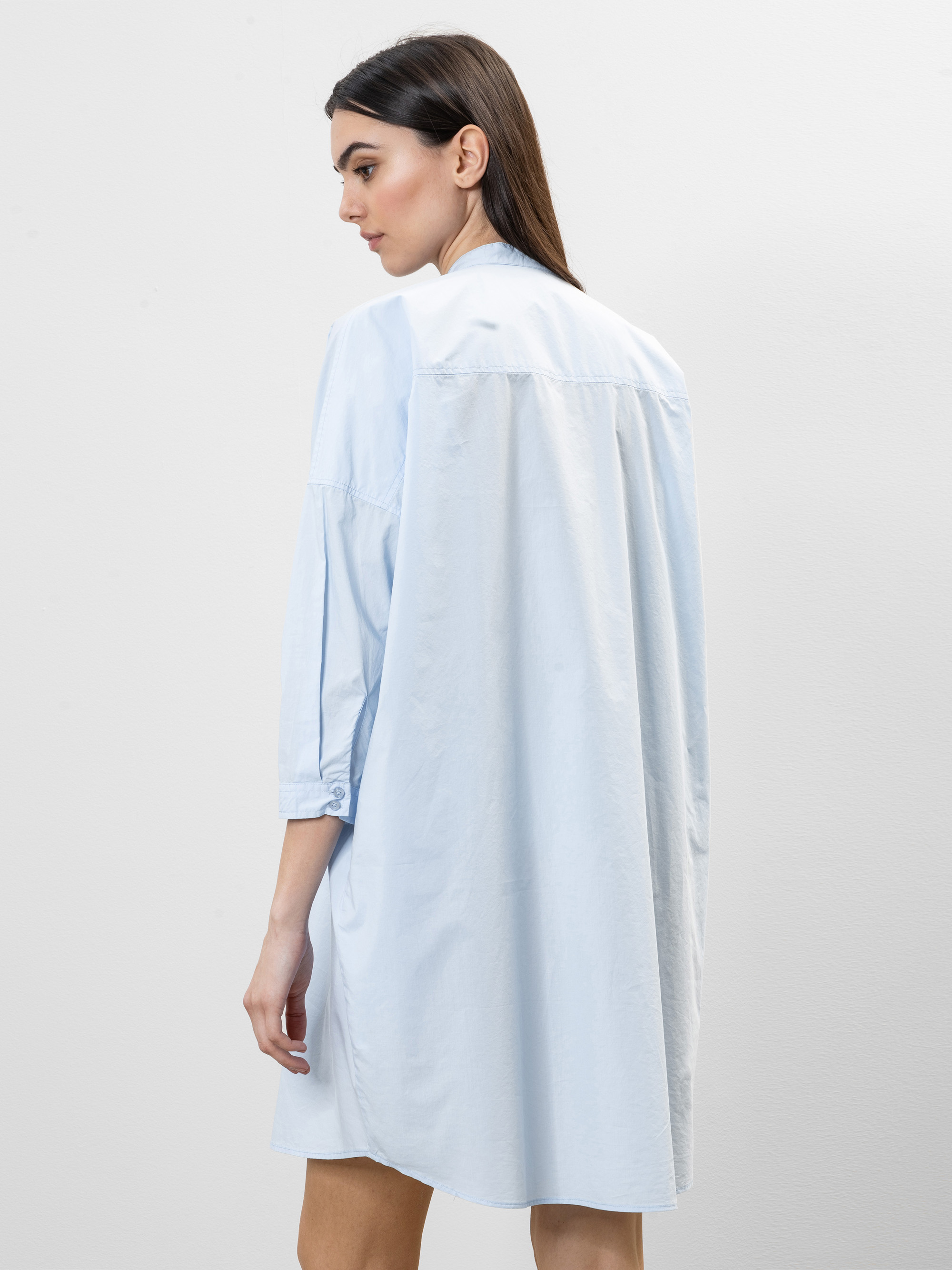 Eine Frau mit langen braunen Haaren trägt ein lockeres, hellblaues Hemdkleid mit Dreiviertelärmeln. Sie steht seitlich und blickt nach unten, der Hintergrund ist einfarbig hell.