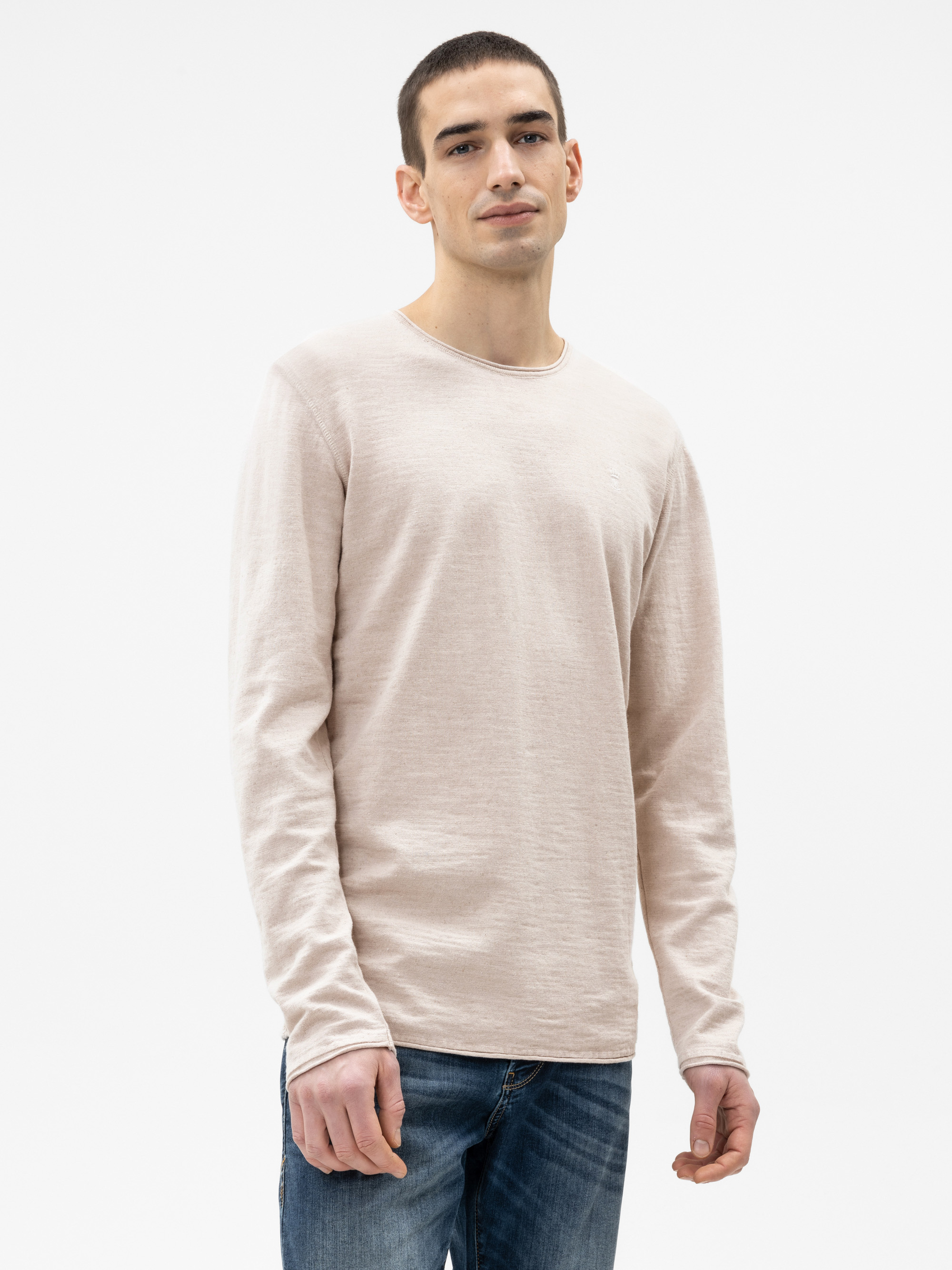 Eine Person steht vor einem schlichten weißen Hintergrund, trägt ein hellbeiges langärmeliges Hemd und eine blaue Jeans und blickt mit neutralem Blick in die Kamera.