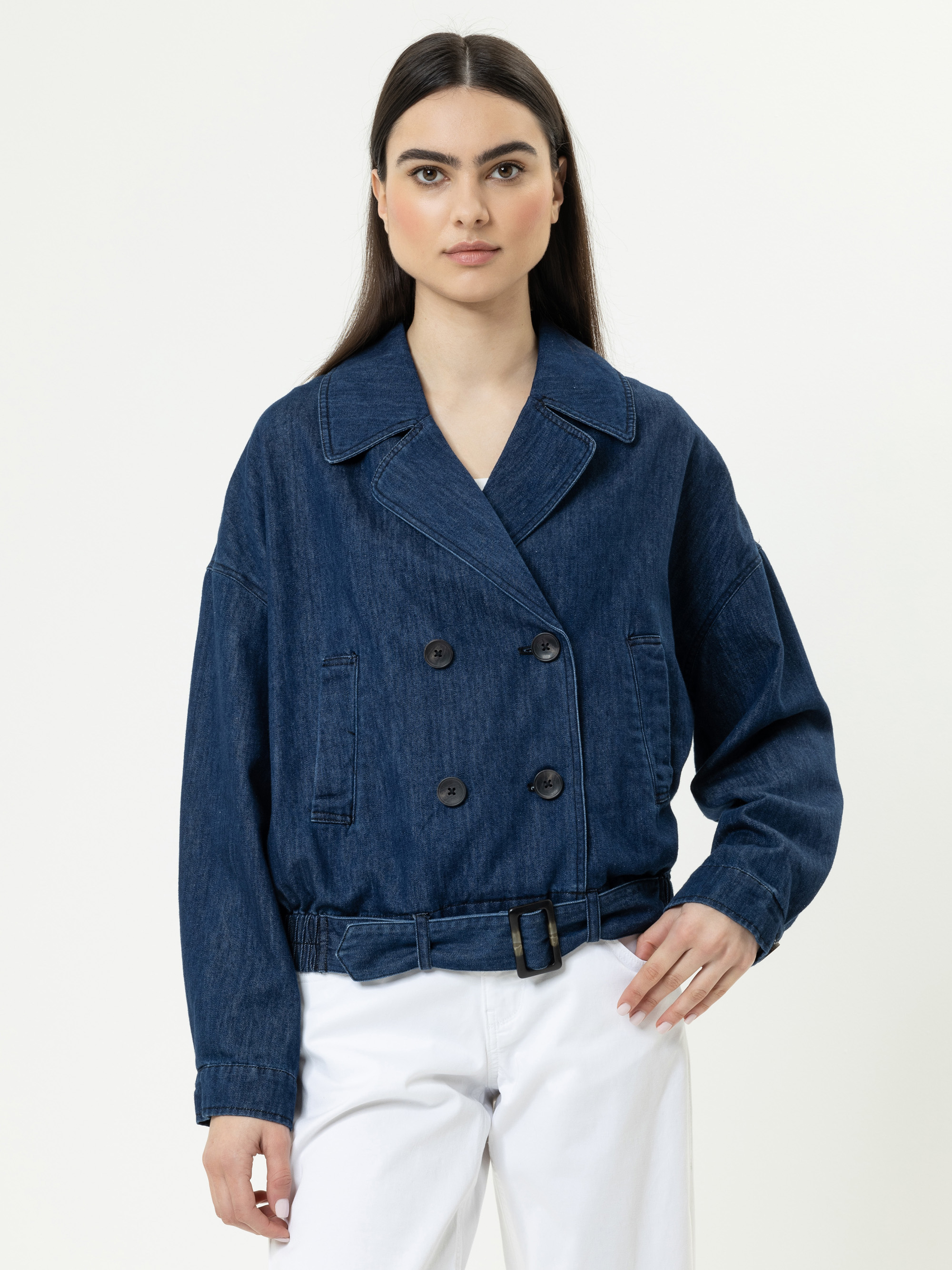Eine Frau mit glattem dunklem Haar trägt eine dunkelblaue doppelreihige Jeansjacke mit Gürtel und eine weiße Hose vor einem schlichten hellen Hintergrund.