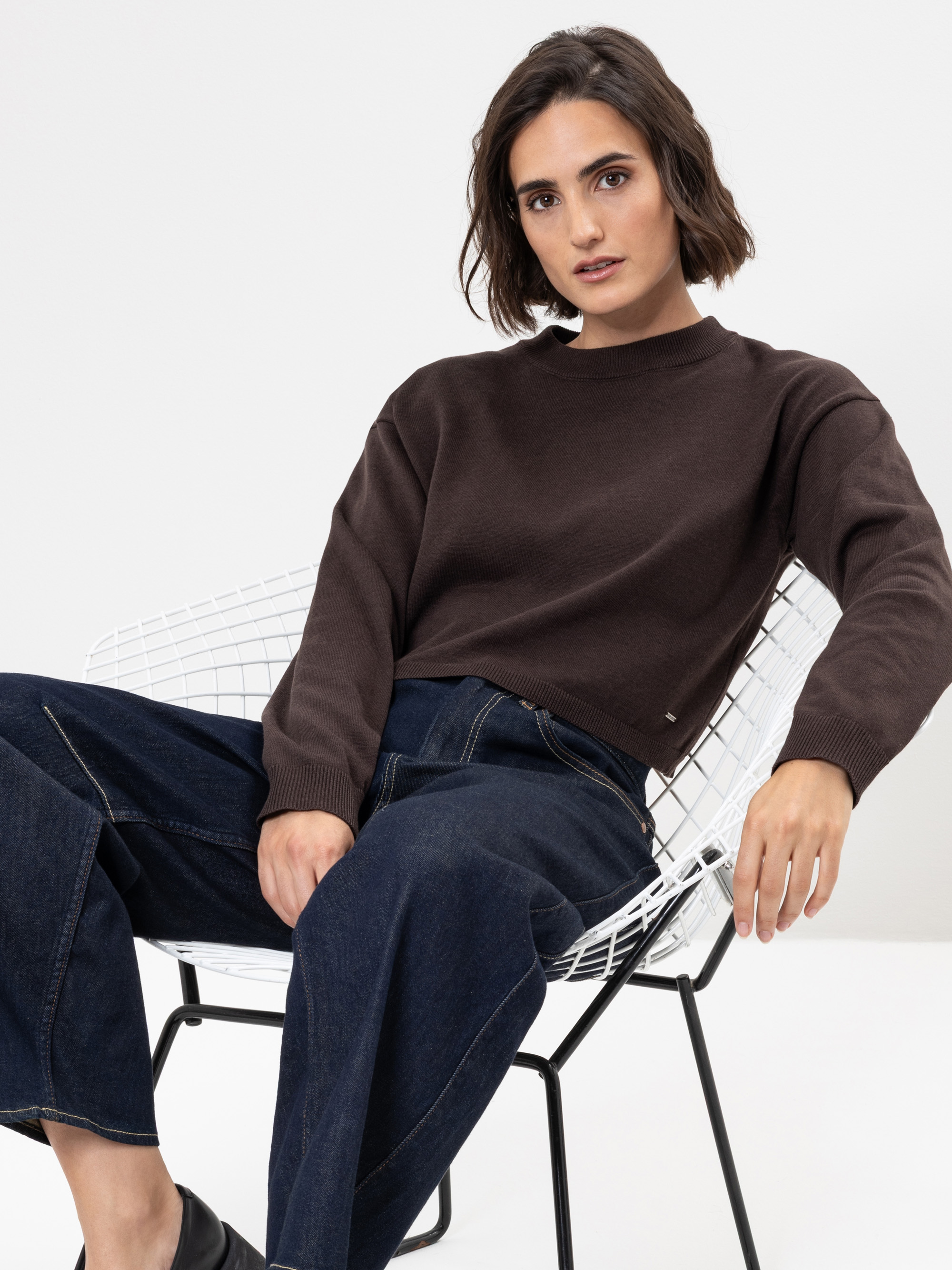 Eine Frau mit kurzen braunen Haaren, die einen dunkelbraunen Pullover und dunkle Jeans mit weitem Bein trägt, sitzt lässig auf einem weißen Drahtstuhl vor einem schlichten weißen Hintergrund.