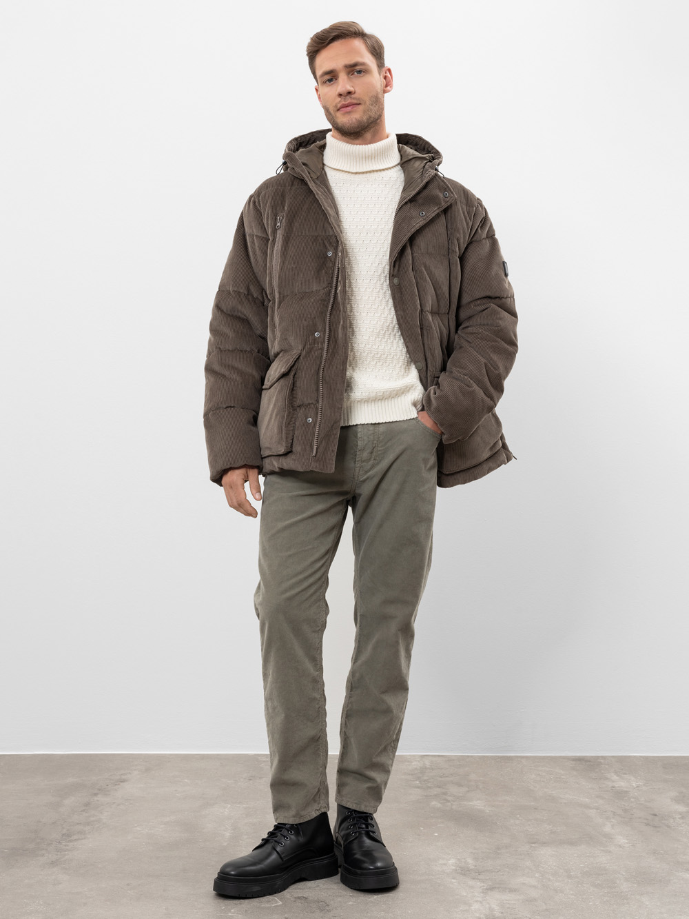 Ein Mann steht in einem Haus und trägt eine braune Pufferjacke, einen weißen Rollkragenpullover, eine olivgrüne Hose und schwarze Schuhe. Er posiert vor einer schlichten weißen Wand und einem grauen Boden.