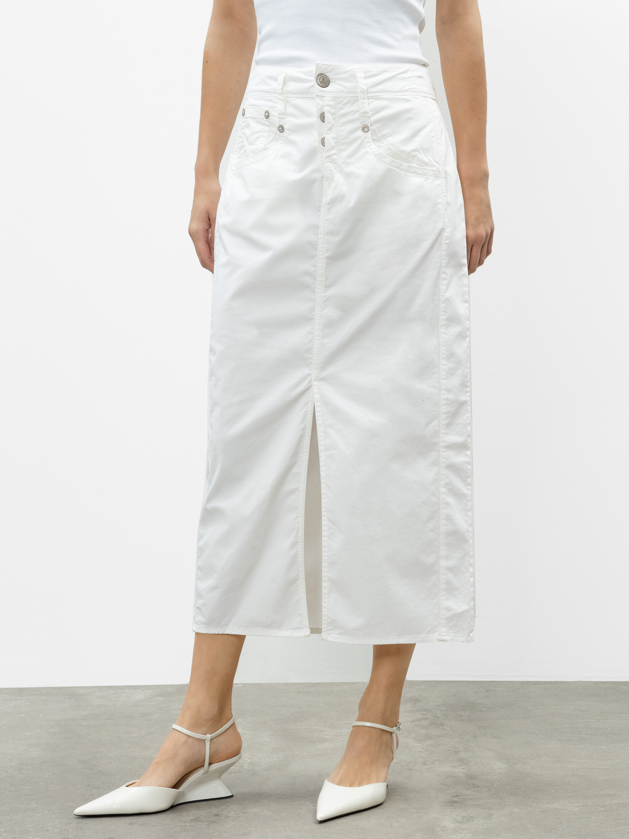 Eine Frau, die einen weißen Midirock mit hoher Taille, Knopfleiste und Schlitz an der Vorderseite trägt, kombiniert mit einem weißen Oberteil und weißen spitzen Absätzen, steht vor einem einfarbigen Hintergrund.