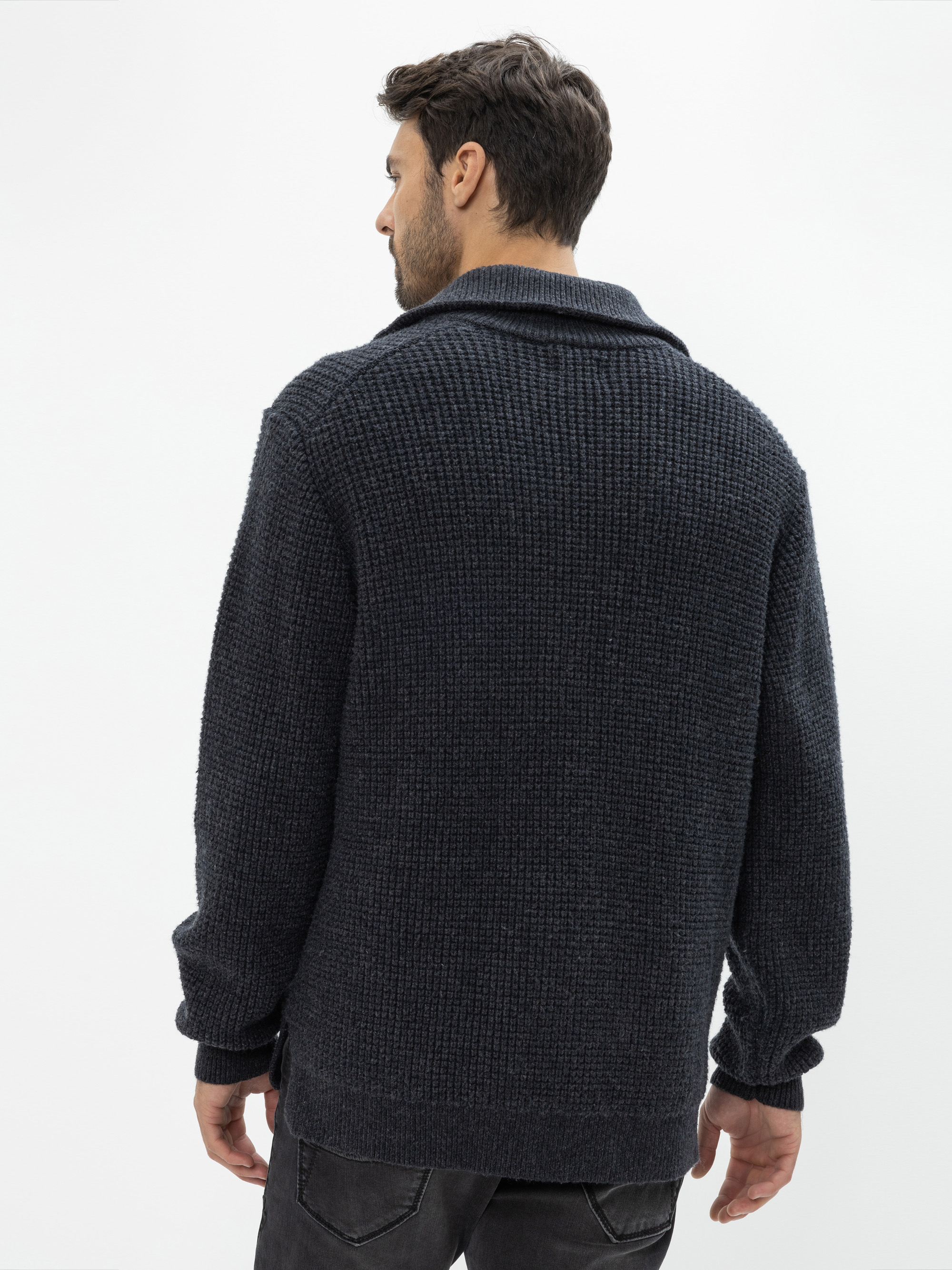 Ein Mann mit kurzen dunklen Haaren wird von hinten gezeigt. Er trägt eine strukturierte dunkelgraue Strickjacke und schwarze Jeans und steht vor einem schlichten weißen Hintergrund.