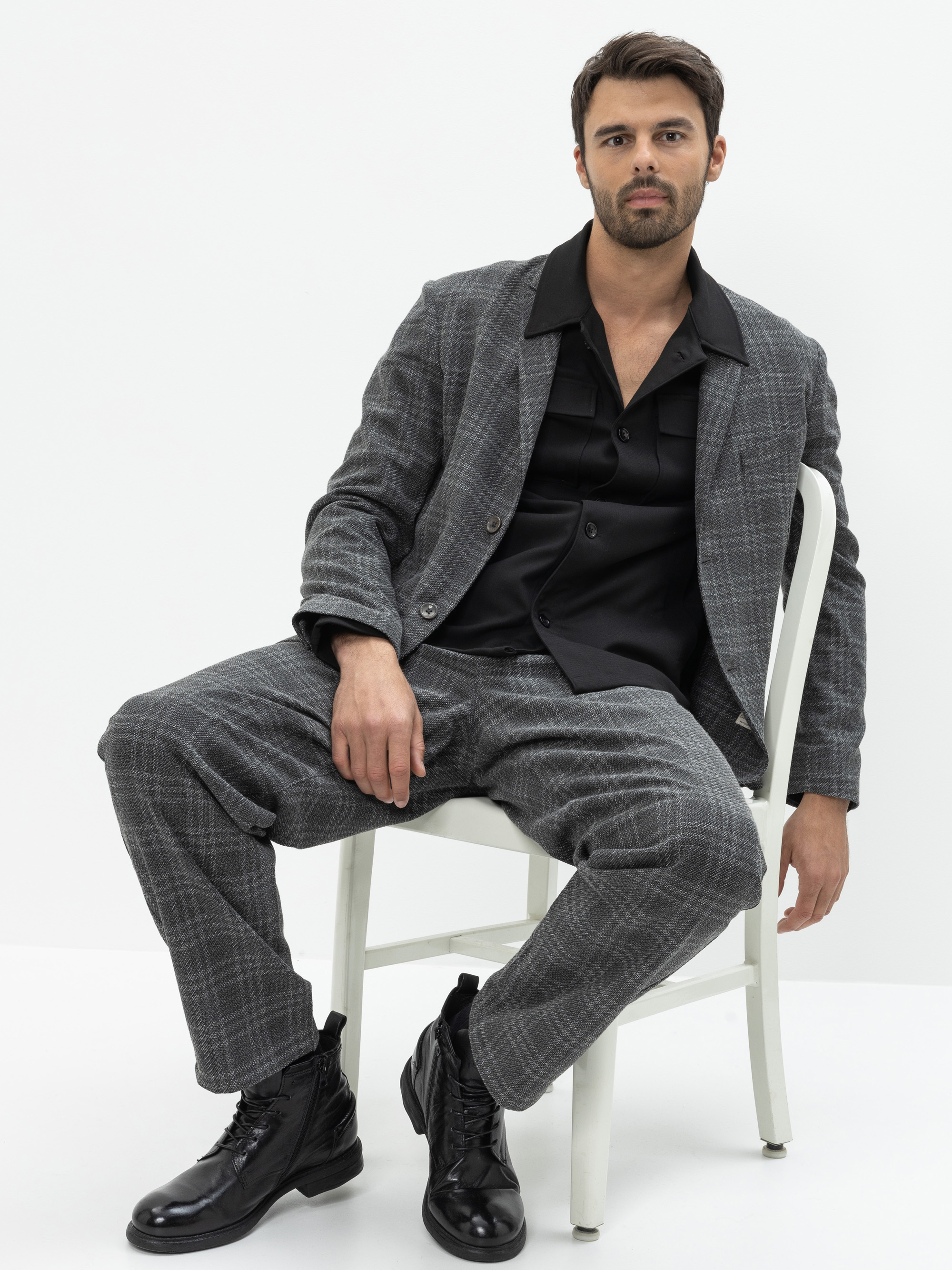 Ein Mann mit kurzen dunklen Haaren und einem Bart sitzt auf einem weißen Stuhl, trägt einen grau karierten Anzug, ein schwarzes Hemd und schwarze Stiefel vor einem schlichten weißen Hintergrund.