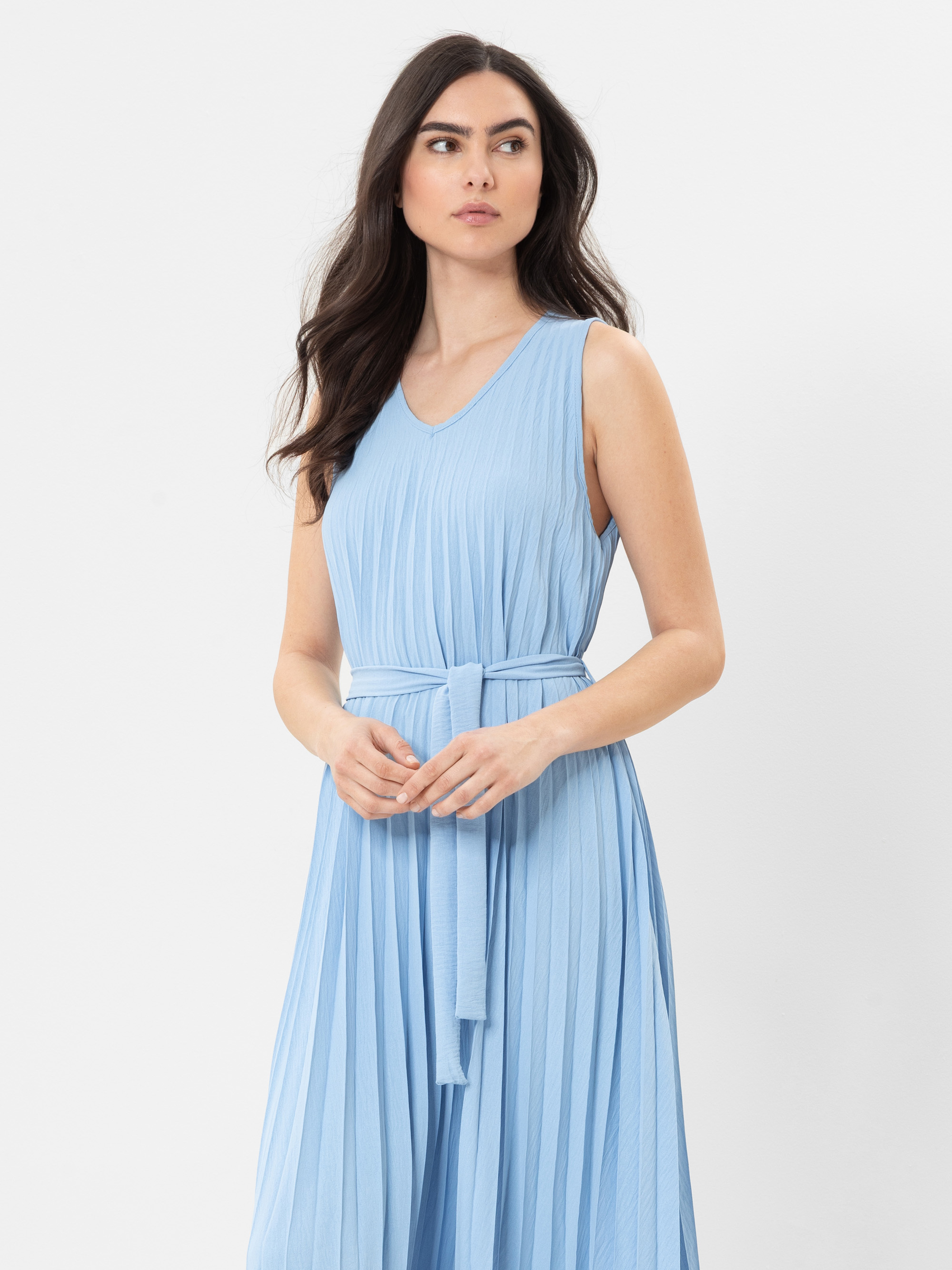 Eine Frau mit langem, dunklem Haar trägt ein ärmelloses, hellblaues, plissiertes Kleid mit passendem Gürtel und steht vor einem schlichten weißen Hintergrund.