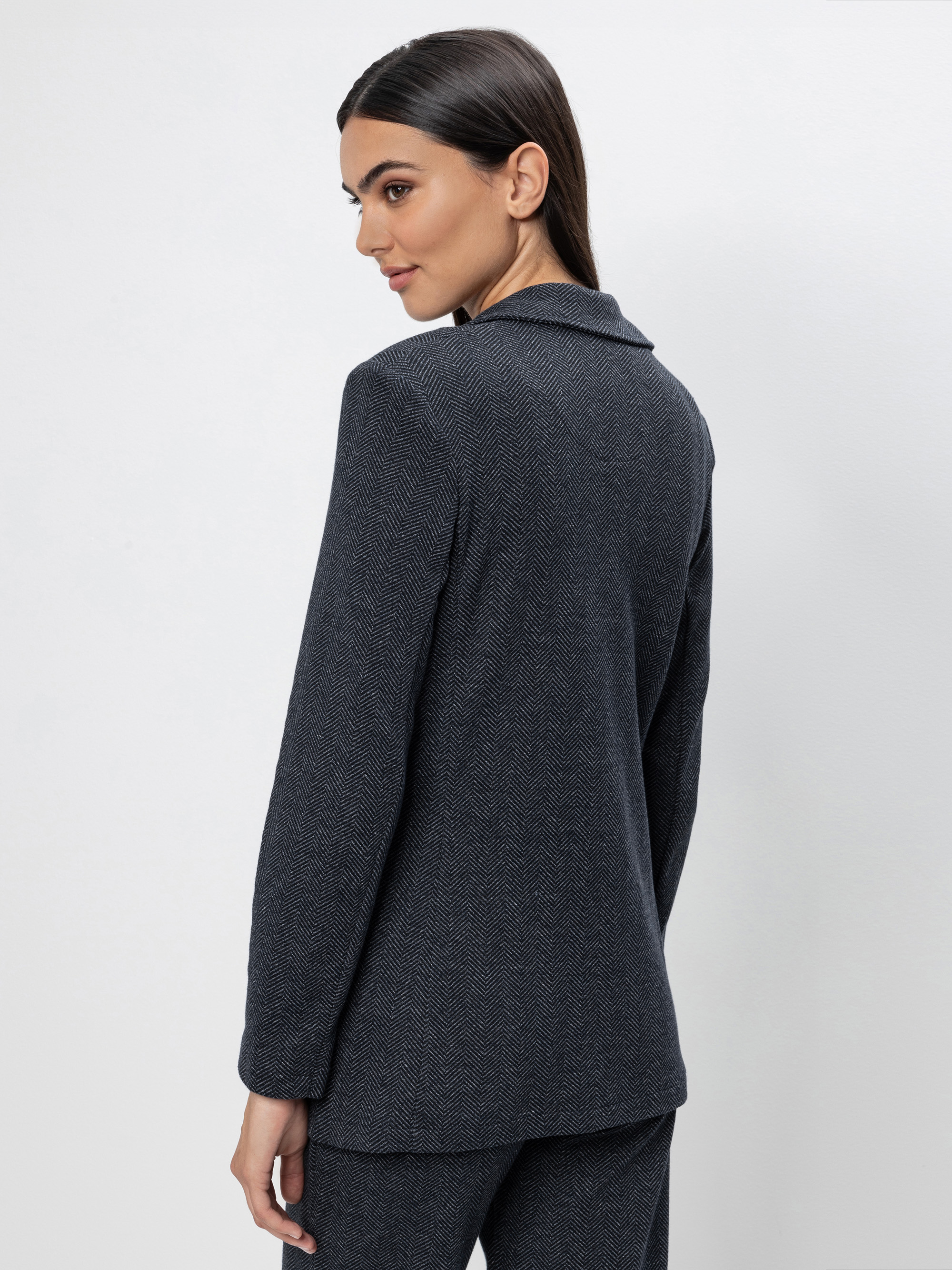 Eine Frau mit glattem dunklem Haar trägt einen dunklen strukturierten Blazer und eine dazu passende Hose. Sie steht leicht zur Seite gedreht und schaut über ihre Schulter vor einem schlichten hellgrauen Hintergrund.