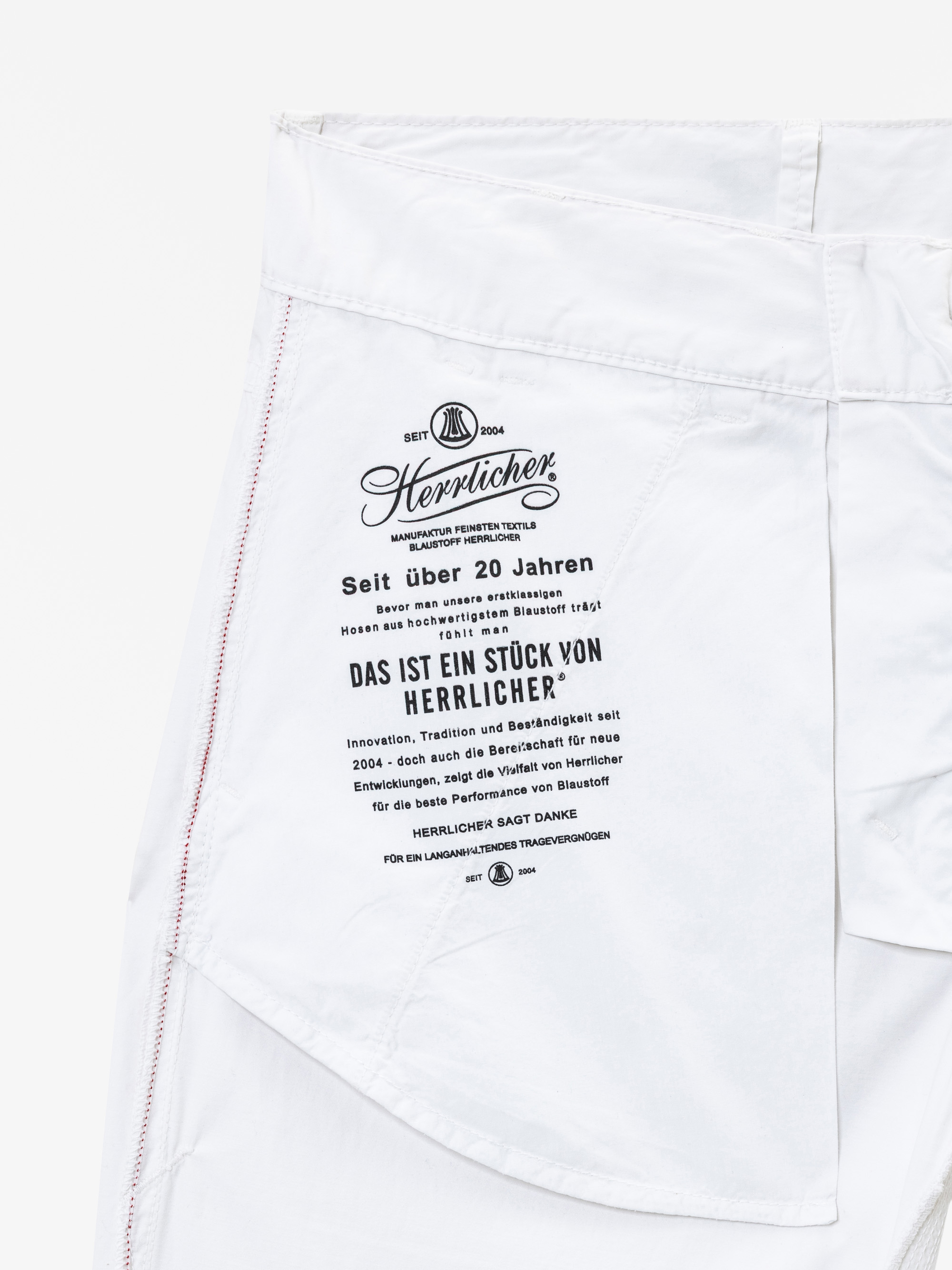 Nahaufnahme einer weißen Hose, die eine Innentasche mit schwarzem Text in deutscher Sprache zeigt, darunter der Markenname "Herrlicher" und eine Botschaft über die Geschichte und die Werte des Unternehmens. An der Naht sind rote Nähte zu sehen.