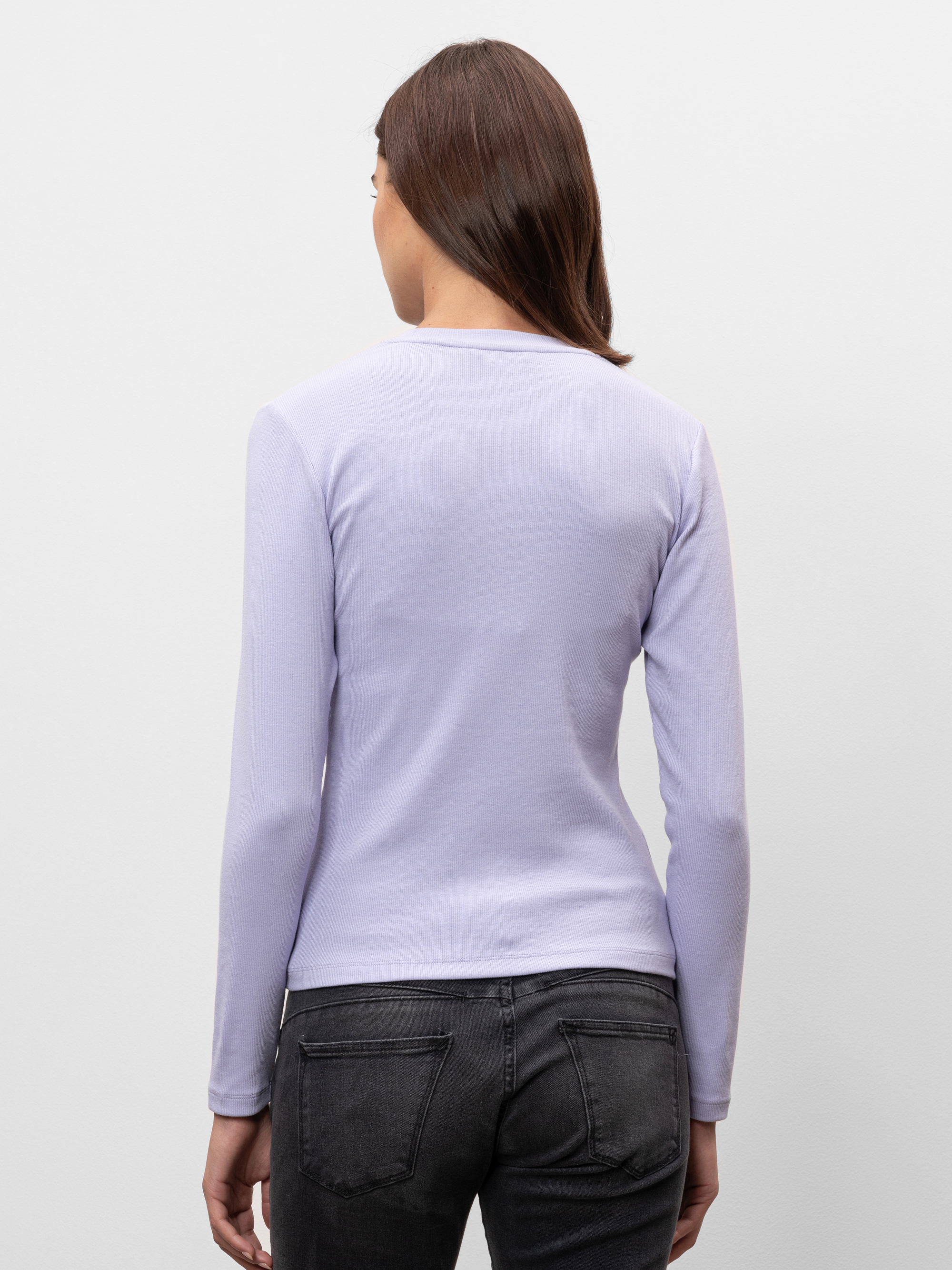 Eine Frau mit glattem braunem Haar steht mit dem Rücken zur Kamera, trägt ein hellviolettes langärmeliges Hemd und dunkelgraue Jeans vor einem schlichten, hellen Hintergrund.