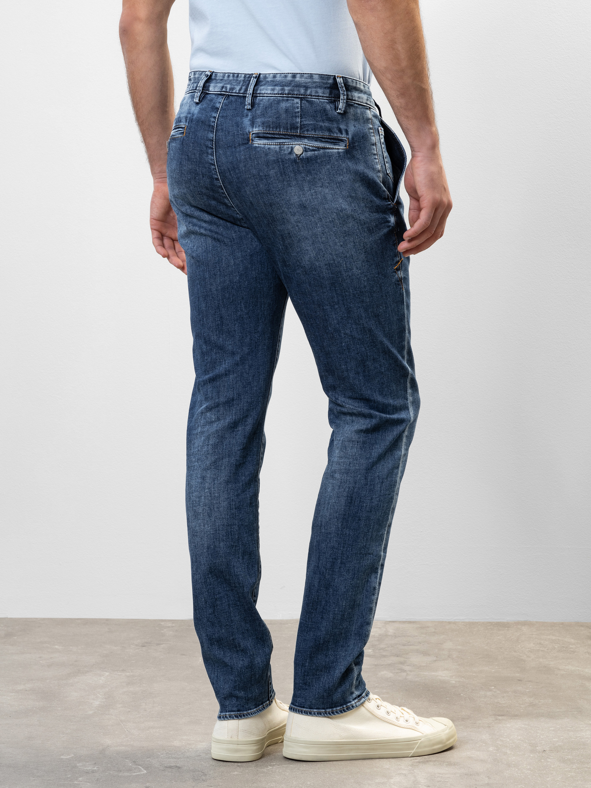 Ein Mann steht auf einem Betonboden und blickt von der Kamera weg. Er trägt eine blaue Slim-Fit-Jeans, ein hellblaues Hemd, das in die Hose gesteckt ist, und weiße Turnschuhe. Der Hintergrund ist eine schlichte hellgraue Wand.