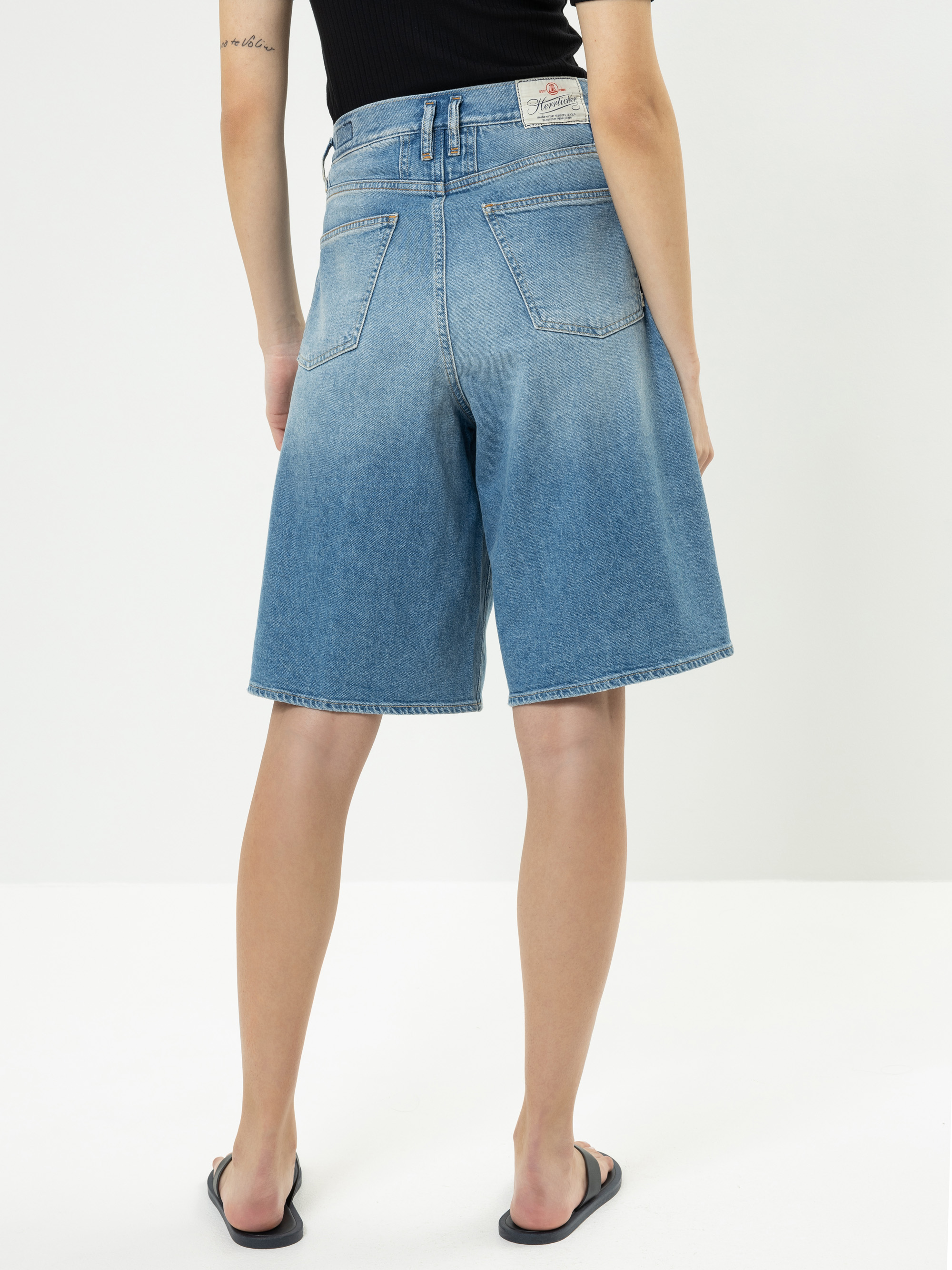 Eine Person, die ein schwarzes ärmelloses Oberteil, blaue knielange Jeans-Shorts mit hoher Taille und schwarze Flip-Flops trägt, steht mit dem Gesicht nach unten vor einem schlichten weißen Hintergrund.
