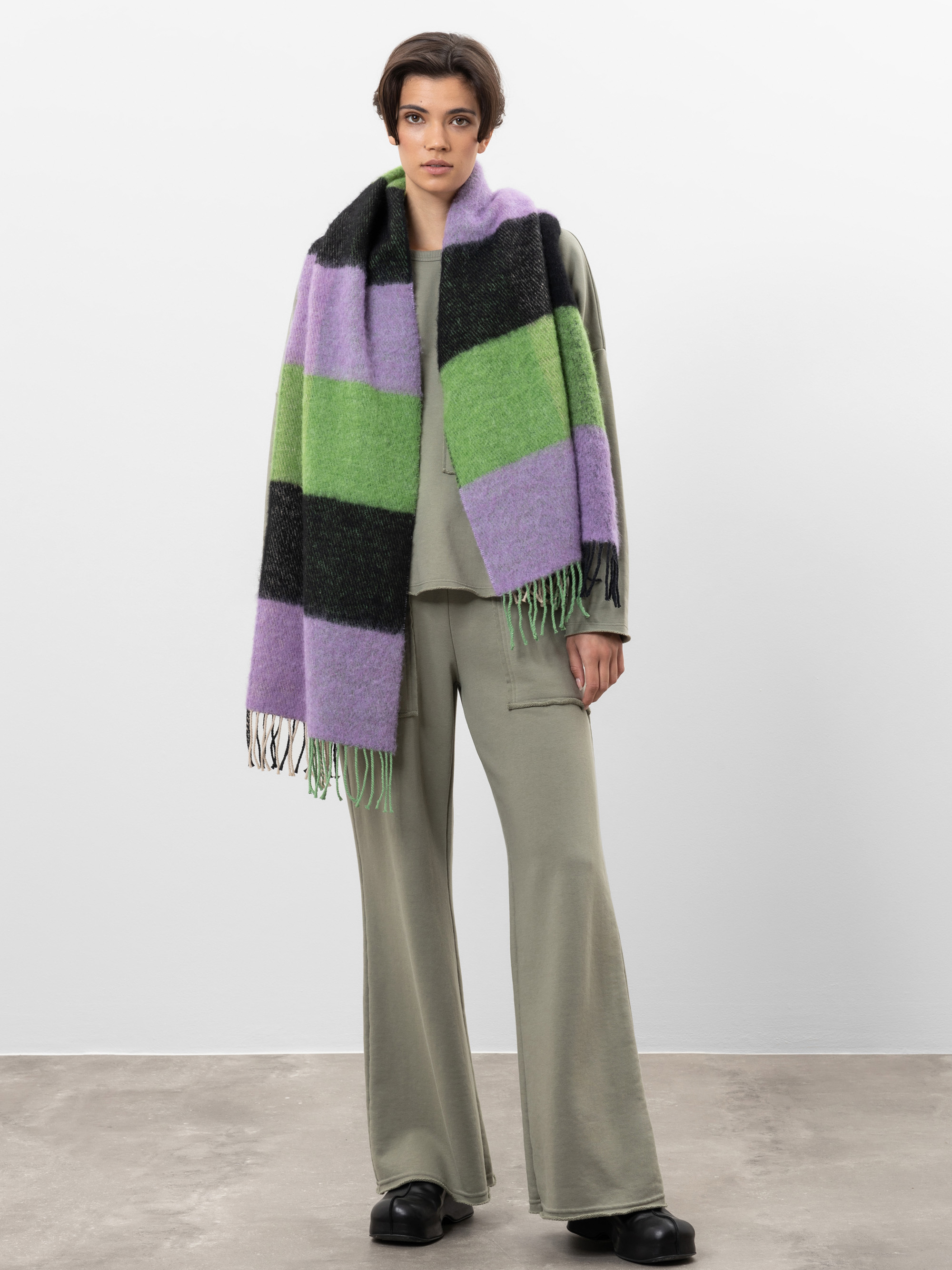 Eine Person, die ein salbeigrünes Lounge-Set mit Schlaghosen, schwarzen Schuhen und einem großen, dicken Schal mit breiten grünen, lila und schwarzen Streifen trägt, steht vor einem schlichten weißen Hintergrund.