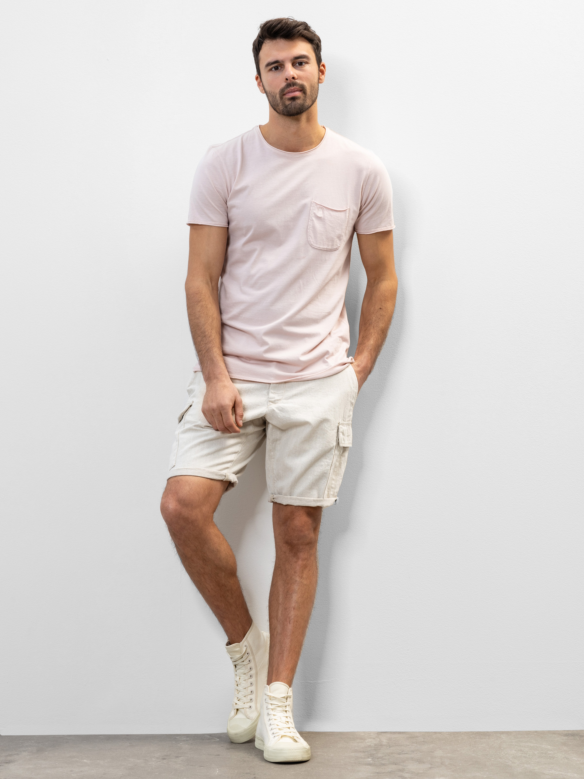 Ein Mann steht an einer weißen Wand und trägt ein hellrosa T-Shirt mit Brusttasche, beigefarbene Cargoshorts und cremefarbene High-Top-Turnschuhe. Er hat kurzes braunes Haar und einen Bart, eine Hand steckt in seiner Tasche.