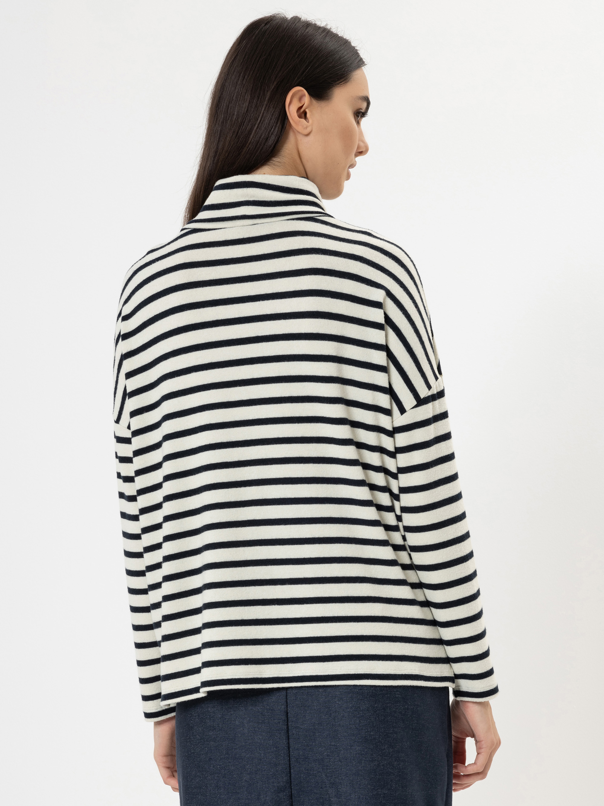 Eine Frau mit langen dunklen Haaren wird von hinten gezeigt. Sie trägt ein locker sitzendes weißes Oberteil mit schwarzen Querstreifen und eine dunkle Hose vor einem schlichten weißen Hintergrund.