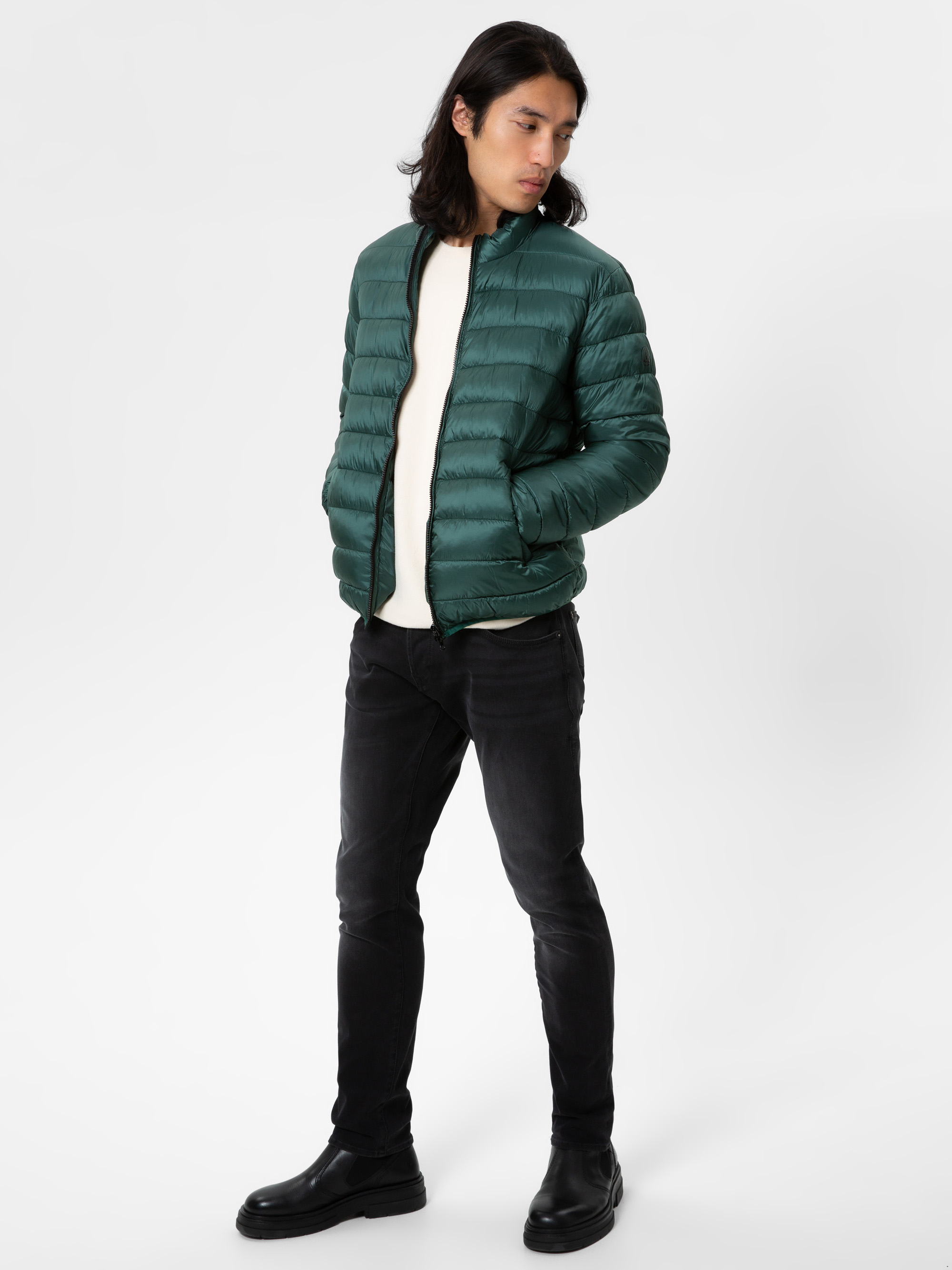 Eine Person mit langen dunklen Haaren trägt eine grüne Pufferjacke über einem weißen Pullover, schwarze Jeans und schwarze Stiefel, steht mit den Händen in den Jackentaschen und blickt zur Seite vor einem schlichten weißen Hintergrund.