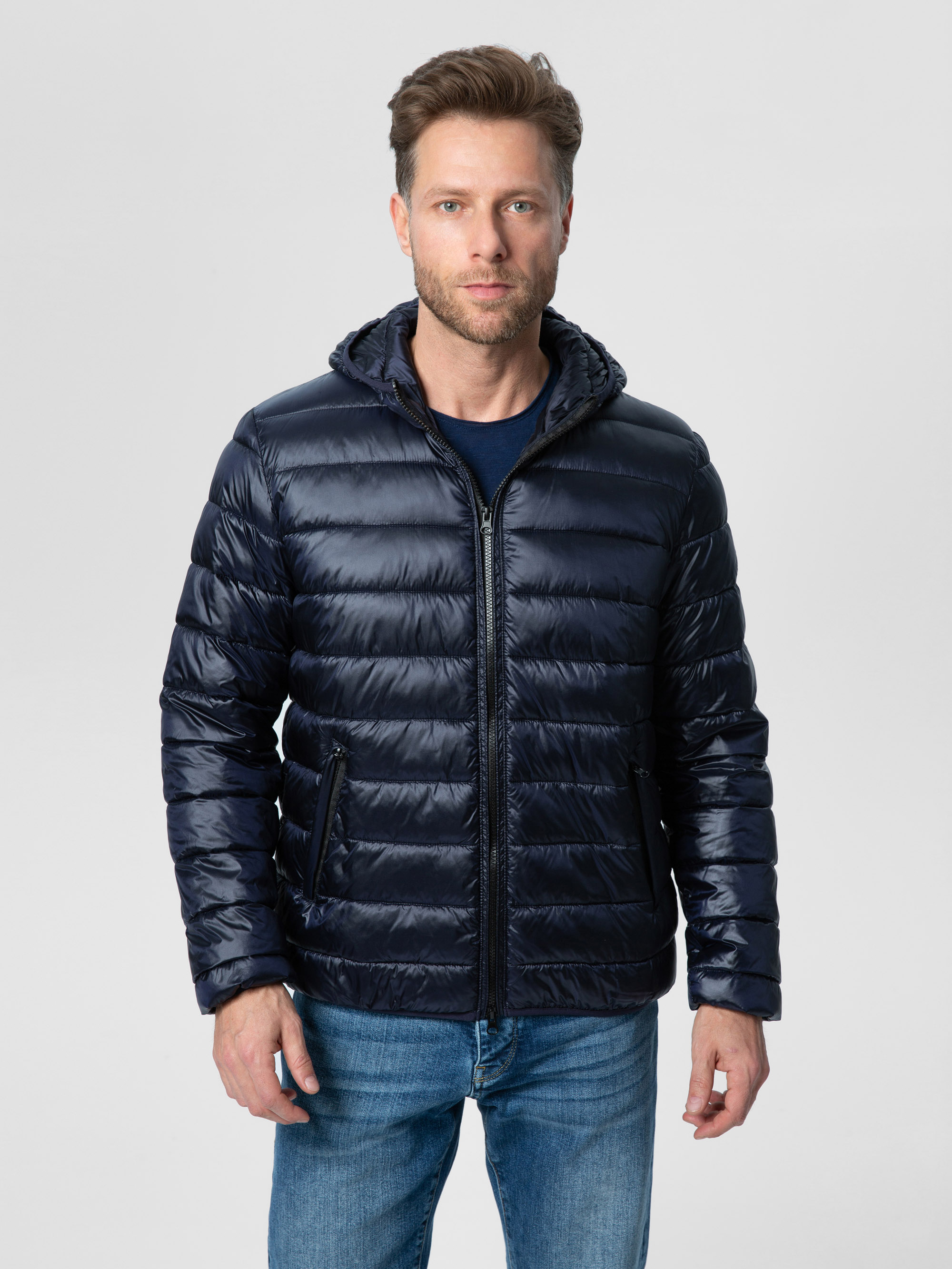 Ein Mann, der eine marineblaue Pufferjacke und blaue Jeans trägt, steht mit dem Gesicht nach vorne vor einem schlichten hellgrauen Hintergrund.