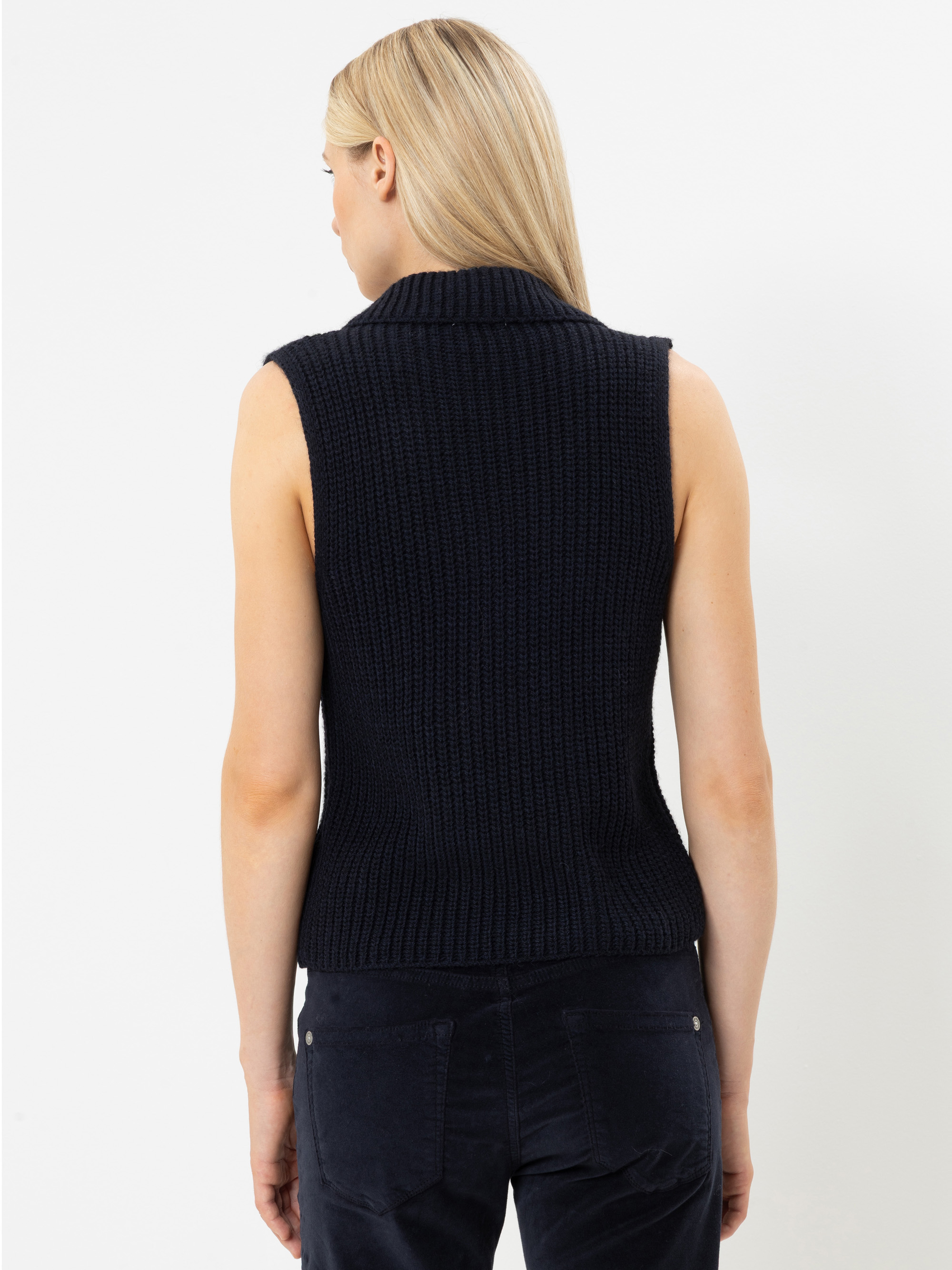 Eine Frau mit langen blonden Haaren ist von hinten zu sehen; sie trägt einen ärmellosen, marineblauen Strickpullover und eine dunkle Hose und steht vor einem schlichten weißen Hintergrund.