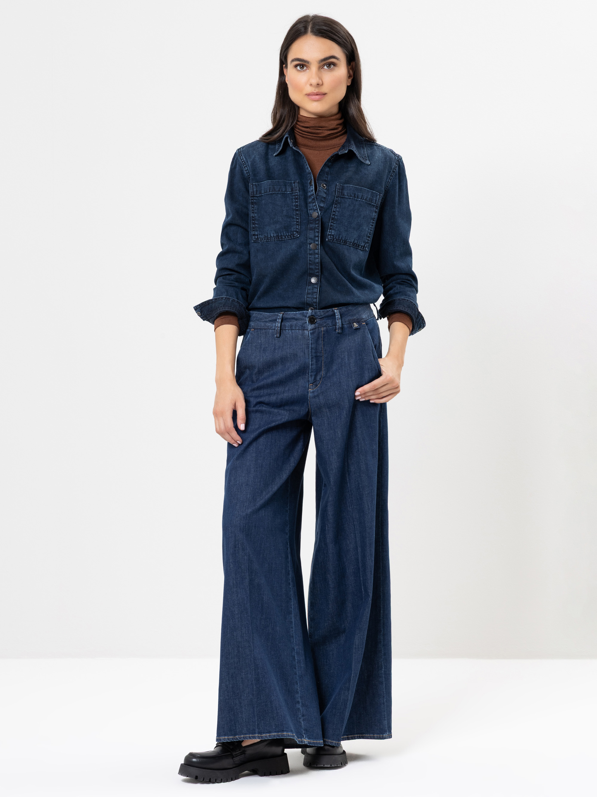 Eine Frau steht vor einem schlichten weißen Hintergrund und trägt ein dunkles Jeanshemd mit Knöpfen und eine Jeanshose mit weitem Bein. Sie hat langes dunkles Haar und schwarze Schuhe. Sie posiert mit einer Hand an der Seite und der anderen auf ihrem O