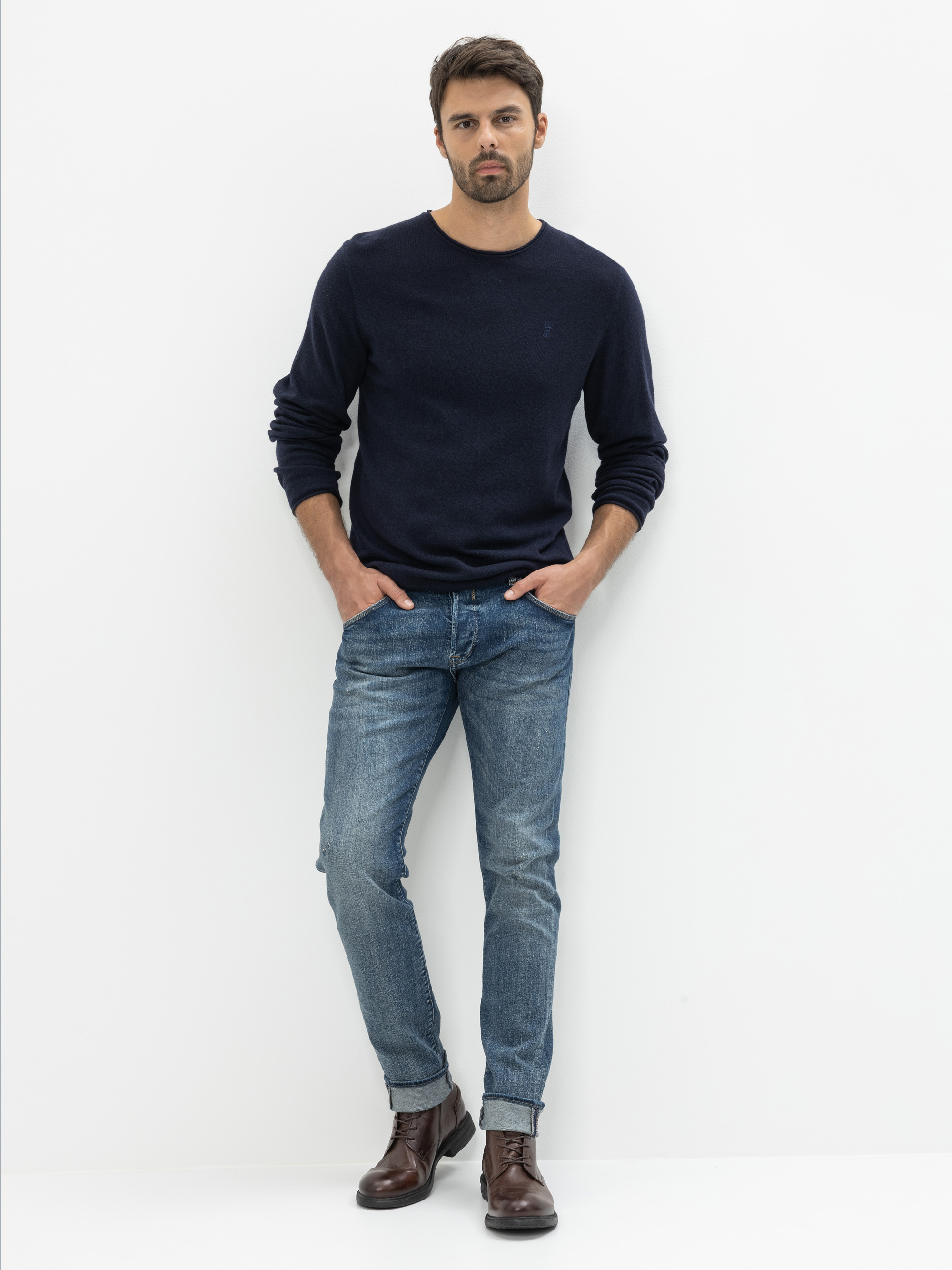 Ein Mann steht vor einem schlichten weißen Hintergrund und trägt einen dunklen marineblauen Pullover, blaue Jeans und braune Lederstiefel. Er hat kurzes dunkles Haar und einen Bart, hat die Hände in den Taschen und schaut direkt in die Kamera.